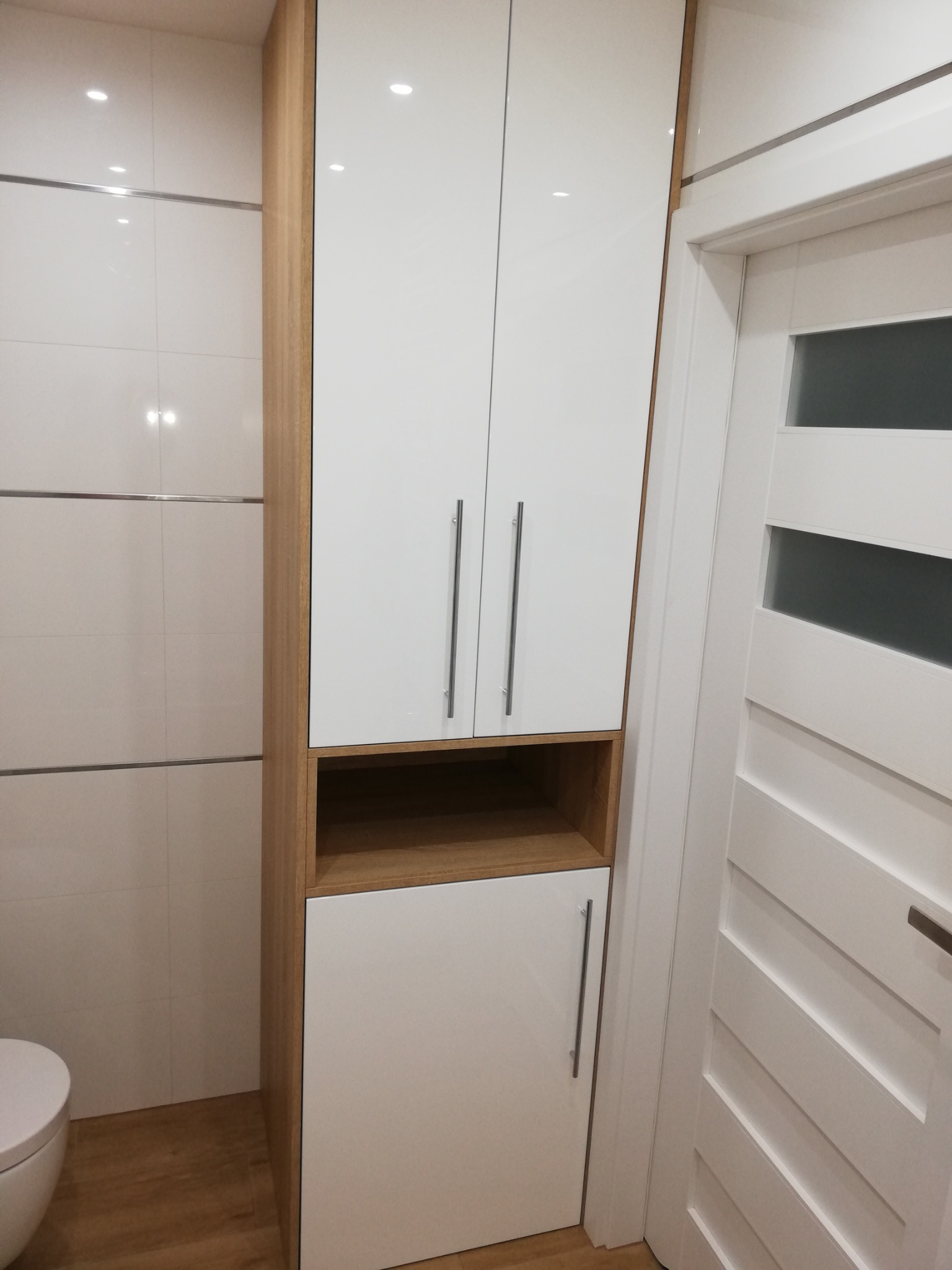 Biała szafka łazienkowa z drewnianymi akcentami, widoczna na tle jasnych płytek i białych drzwi. Minimalistyczny design, pionowe uchwyty, oświetlenie punktowe.