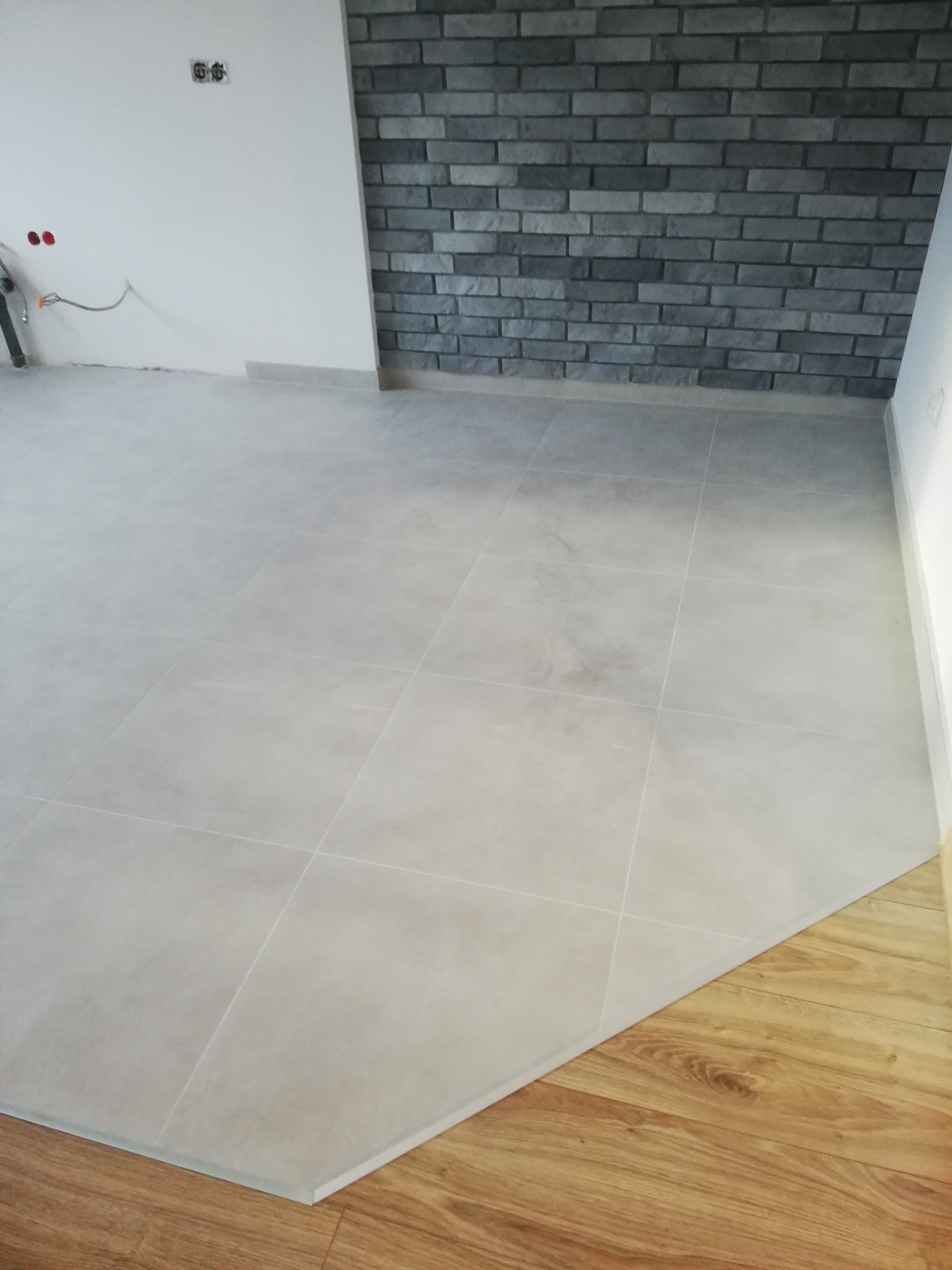 Jasnoszare płytki podłogowe w nowym wnętrzu, z fragmentem drewnianej podłogi i dekoracyjną ścianą z szarej cegły. Widoczne gniazdka elektryczne.