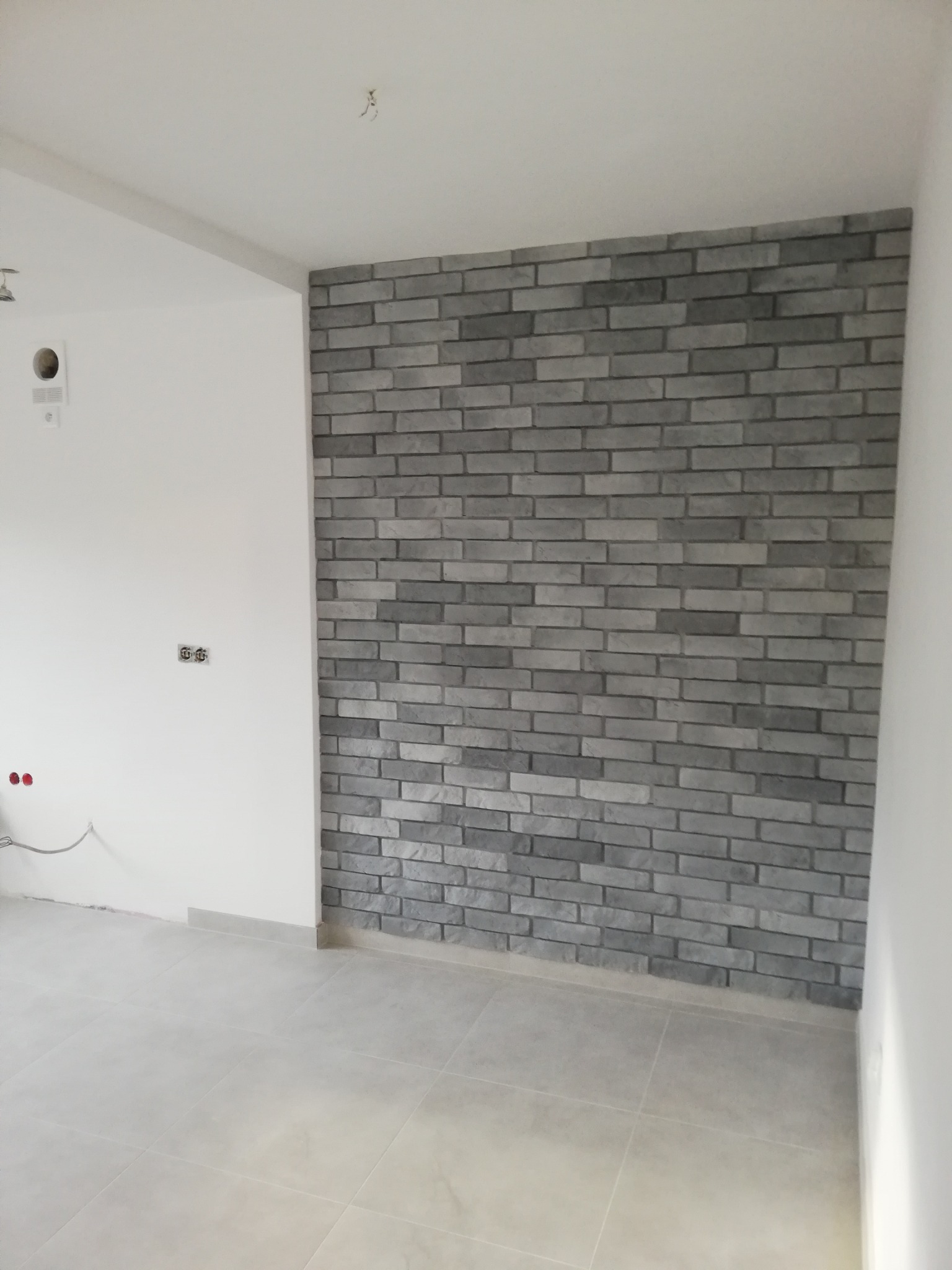 Ściana z szarej cegły dekoracyjnej w nowym wnętrzu. Widoczne gniazdka elektryczne i fragment podłogi z płytek. Minimalistyczny design, surowe wykończenie.
