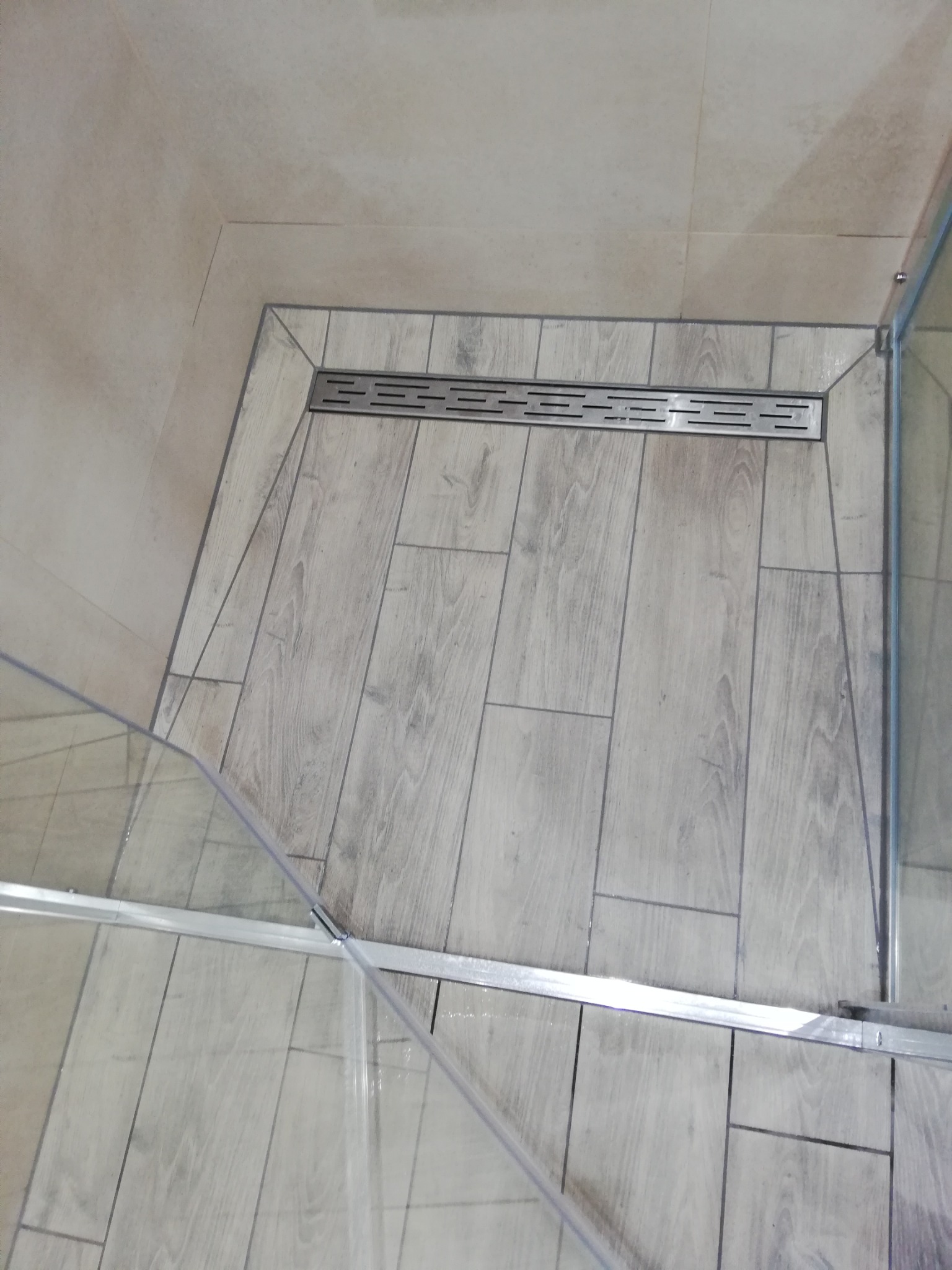 Wykończona strefa prysznicowa z odpływem liniowym i płytkami imitującymi drewno. Fragment szklanej kabiny prysznicowej.