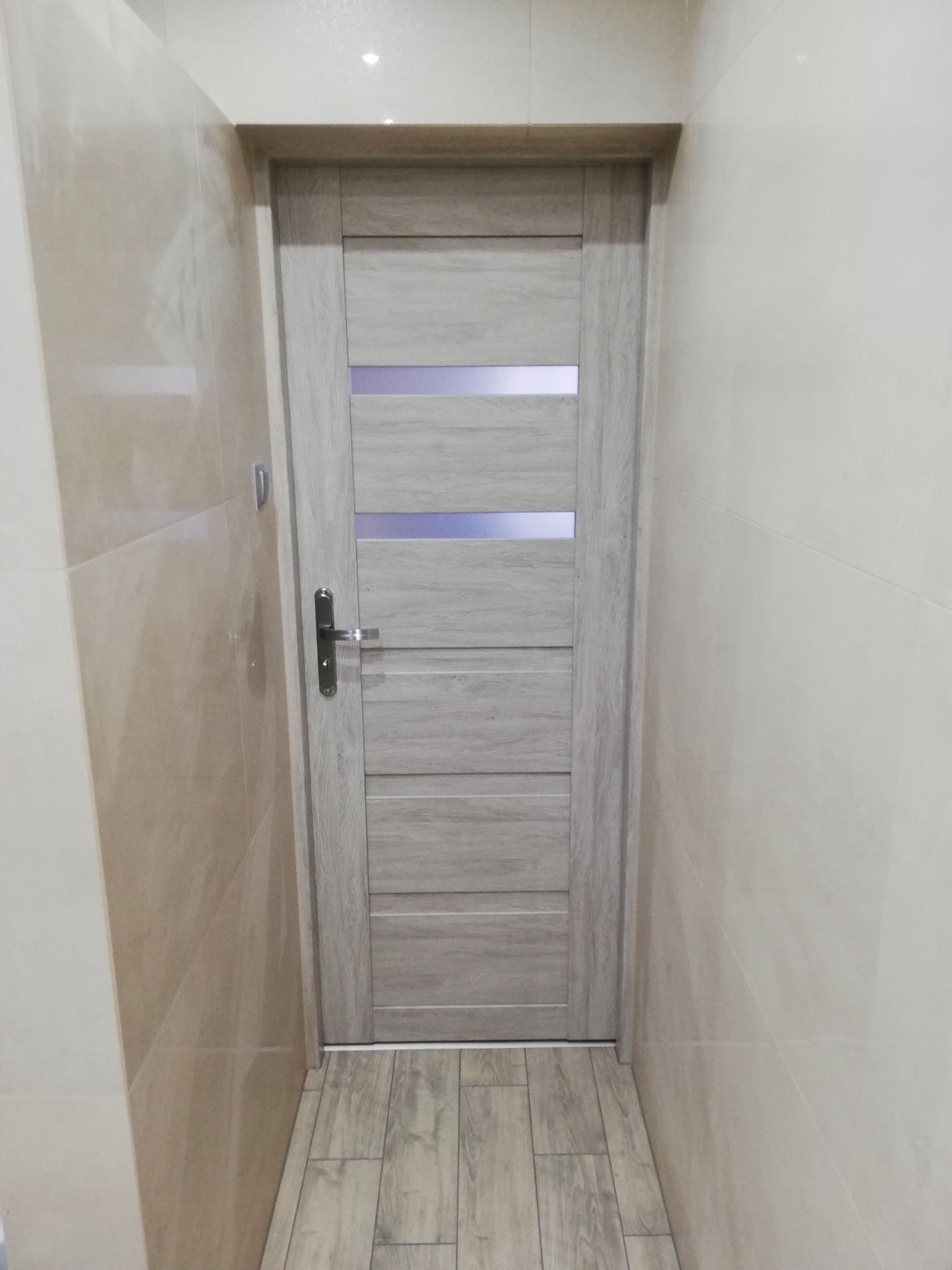 Zamknięte, drewnopodobne drzwi z szybkami w łazience wykończonej beżowymi płytkami i drewnianą podłogą. Widok na fragment ściany i podłogi.