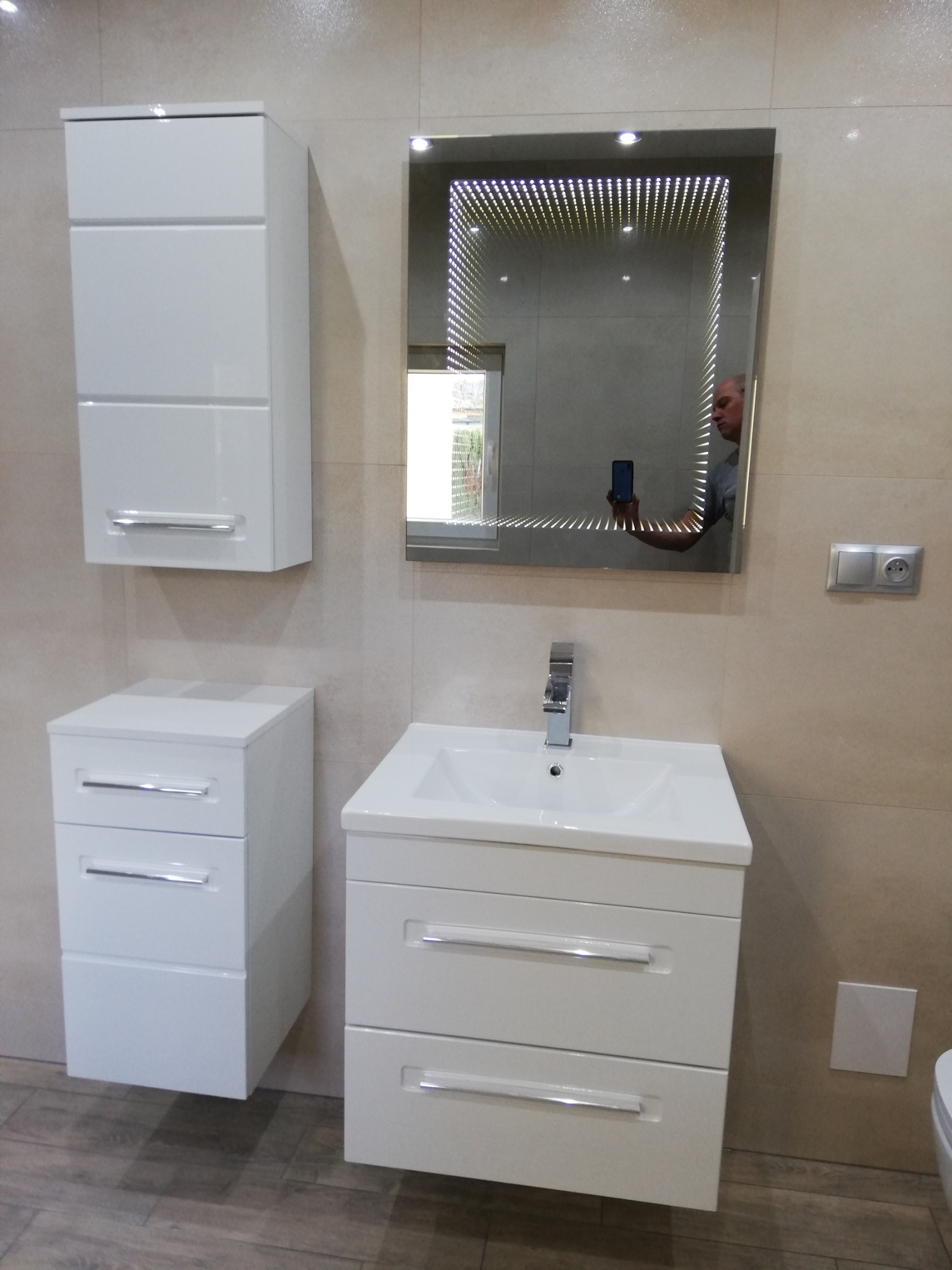 Biała szafka łazienkowa z umywalką i lustrem z efektem nieskończoności, odbicie osoby robiącej zdjęcie. Beżowe płytki na ścianach i drewnopodobna podłoga.