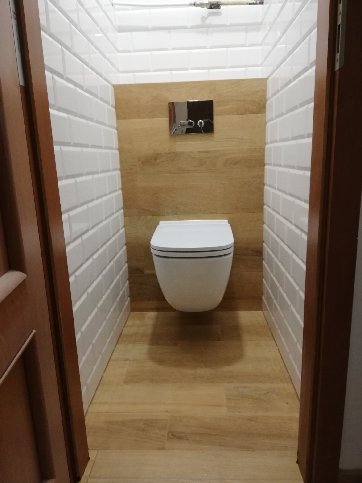 Nowoczesna toaleta w wąskiej łazience z białymi, wypukłymi płytkami na ścianach i drewnopodobną okładziną. Widok zza uchylonych, drewnianych drzwi.