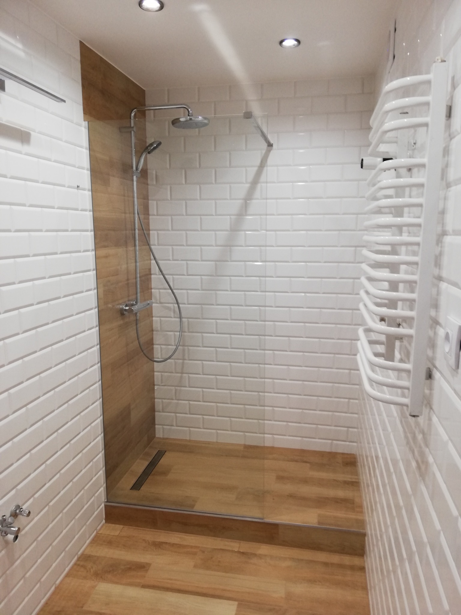 Nowoczesna kabina prysznicowa walk-in z drewnopodobną podłogą i białymi, cegiełkowymi płytkami. Minimalistyczny design, grzejnik drabinkowy.