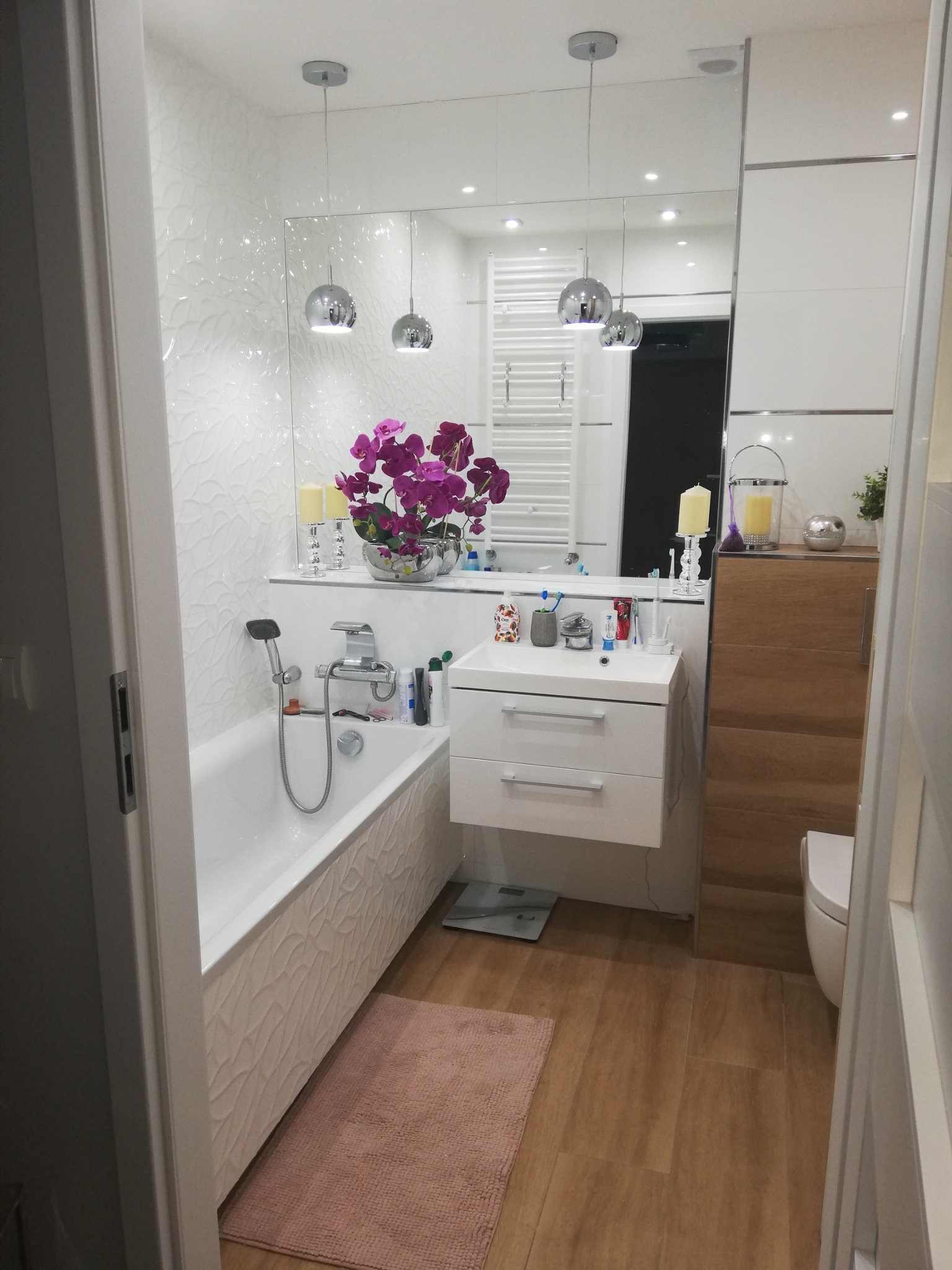 Nowoczesna łazienka z wanną, umywalką z szafką i dużym lustrem. Białe płytki z subtelnym wzorem, drewnopodobna podłoga i fioletowe kwiaty dodają elegancji.