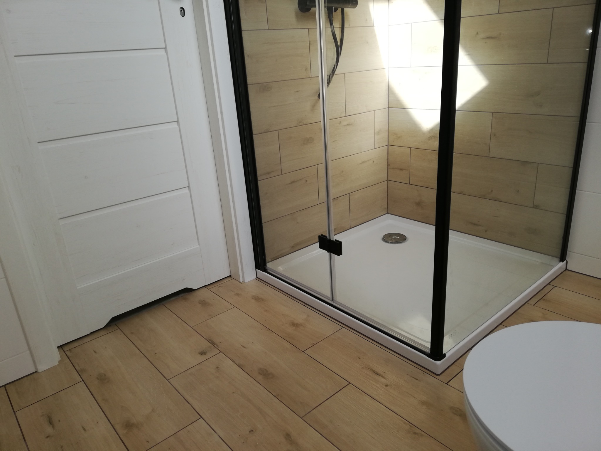 Nowoczesna kabina prysznicowa z czarnymi profilami, jasne płytki imitujące drewno na ścianach i podłodze, białe drzwi i fragment toalety. Minimalistyczny design.