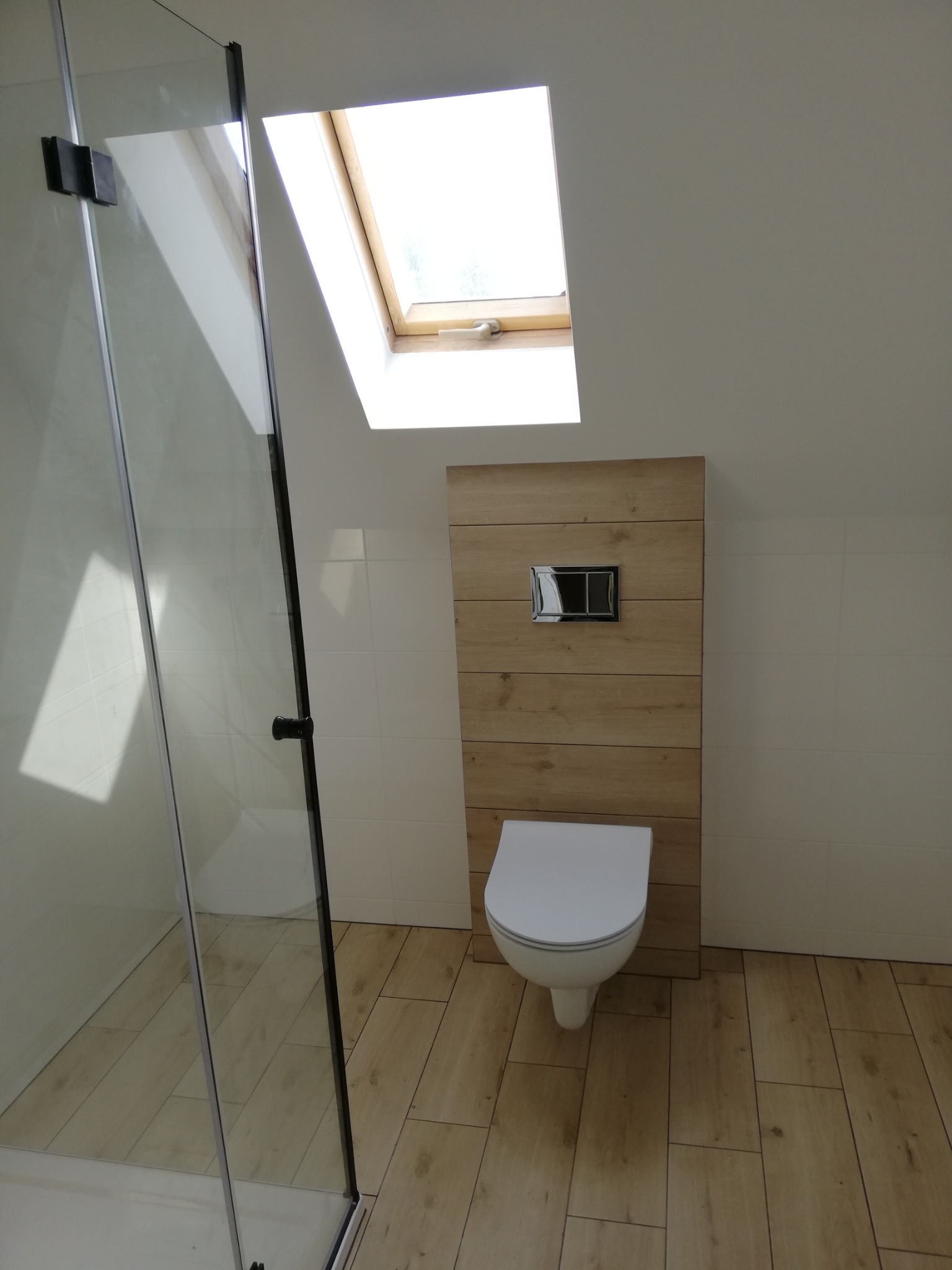 Nowoczesna łazienka na poddaszu z kabiną prysznicową, toaletą podwieszaną na drewnianej ściance i oknem dachowym. Jasne płytki i drewnopodobna podłoga.