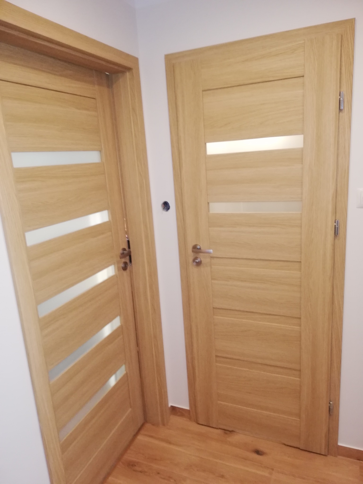 Dwie drewniane drzwi wewnętrzne z przeszkleniami, zamontowane w jasnym pomieszczeniu. Jedne drzwi z ukośnymi szybami, drugie z poziomymi. Widoczna podłoga z paneli.