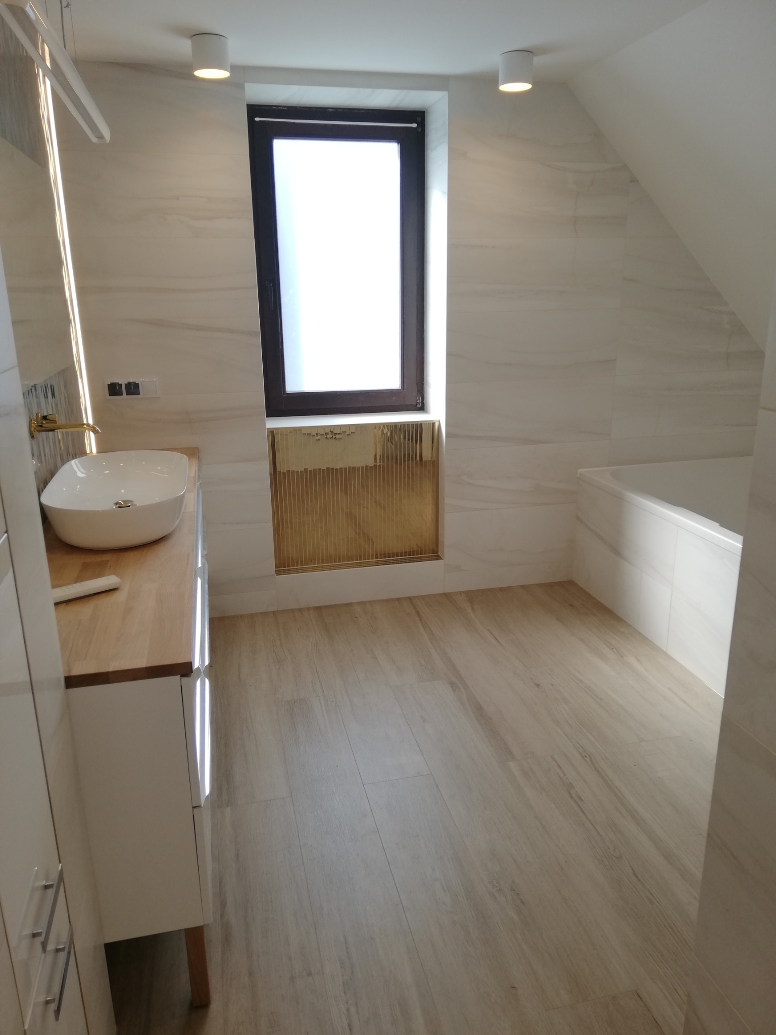 Wykończona łazienka na poddaszu z drewnianą podłogą, umywalką na blacie i oknem z dekoracyjną wnęką. Białe płytki imitujące marmur na ścianach i minimalistyczne oświetlenie.