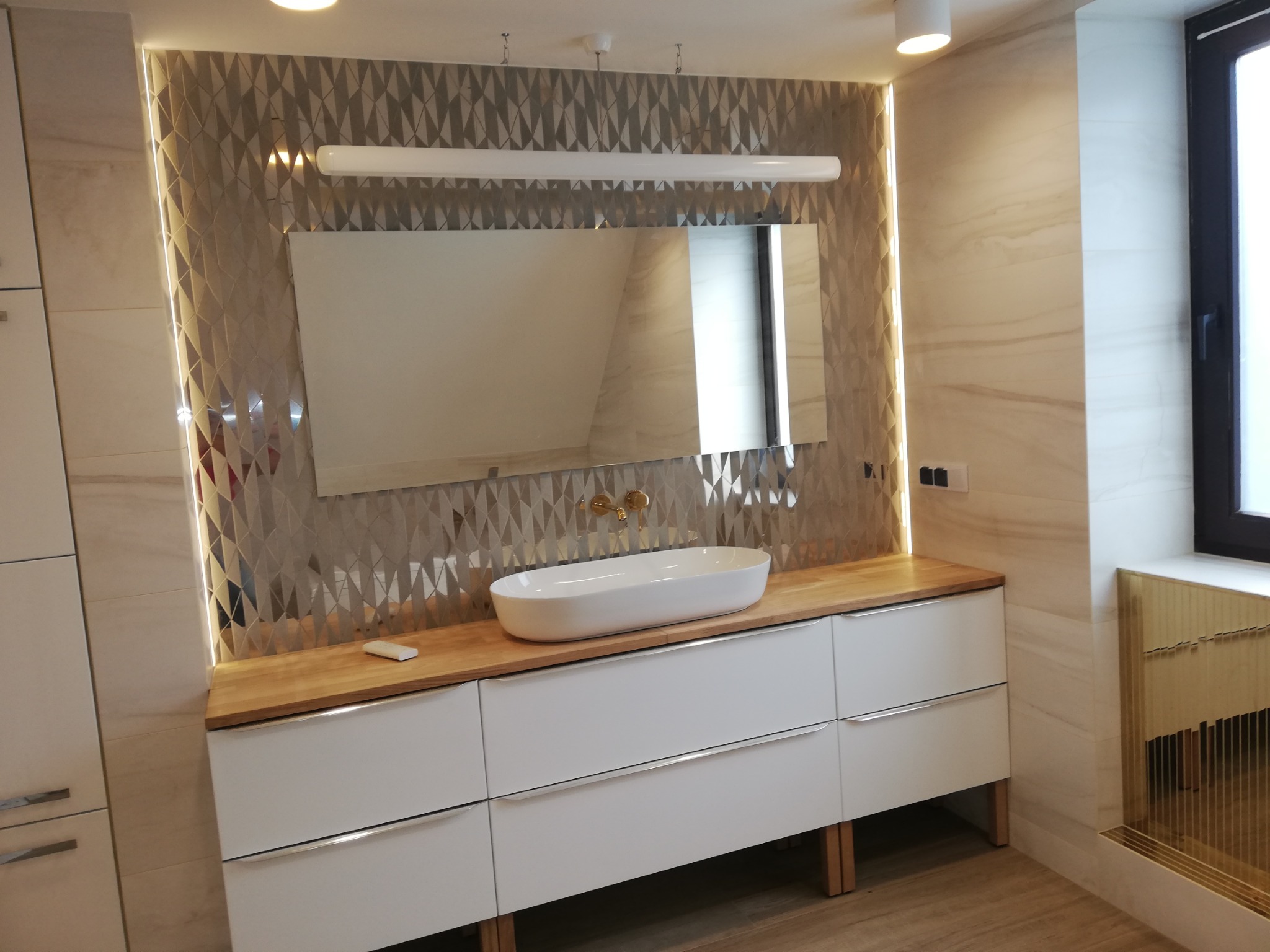 Nowoczesna łazienka z umywalką nablatową, drewnianym blatem i geometryczną mozaiką na ścianie, oświetlona listwą LED. Minimalistyczny design z białymi szafkami.