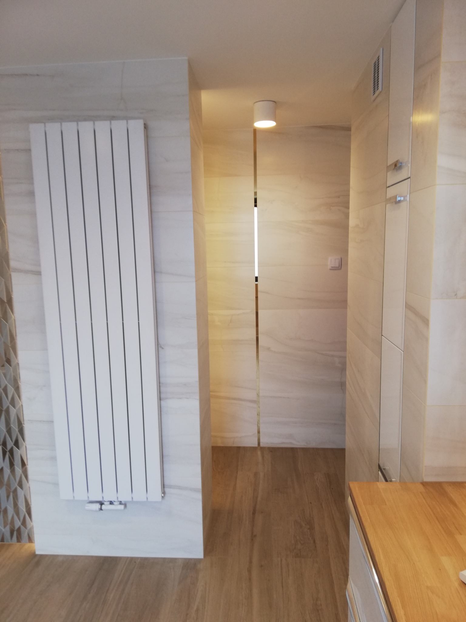 Nowoczesna łazienka z białym grzejnikiem panelowym, marmurowymi płytkami i drewnianą podłogą. Widok na oświetlone przejście do dalszej części pomieszczenia.