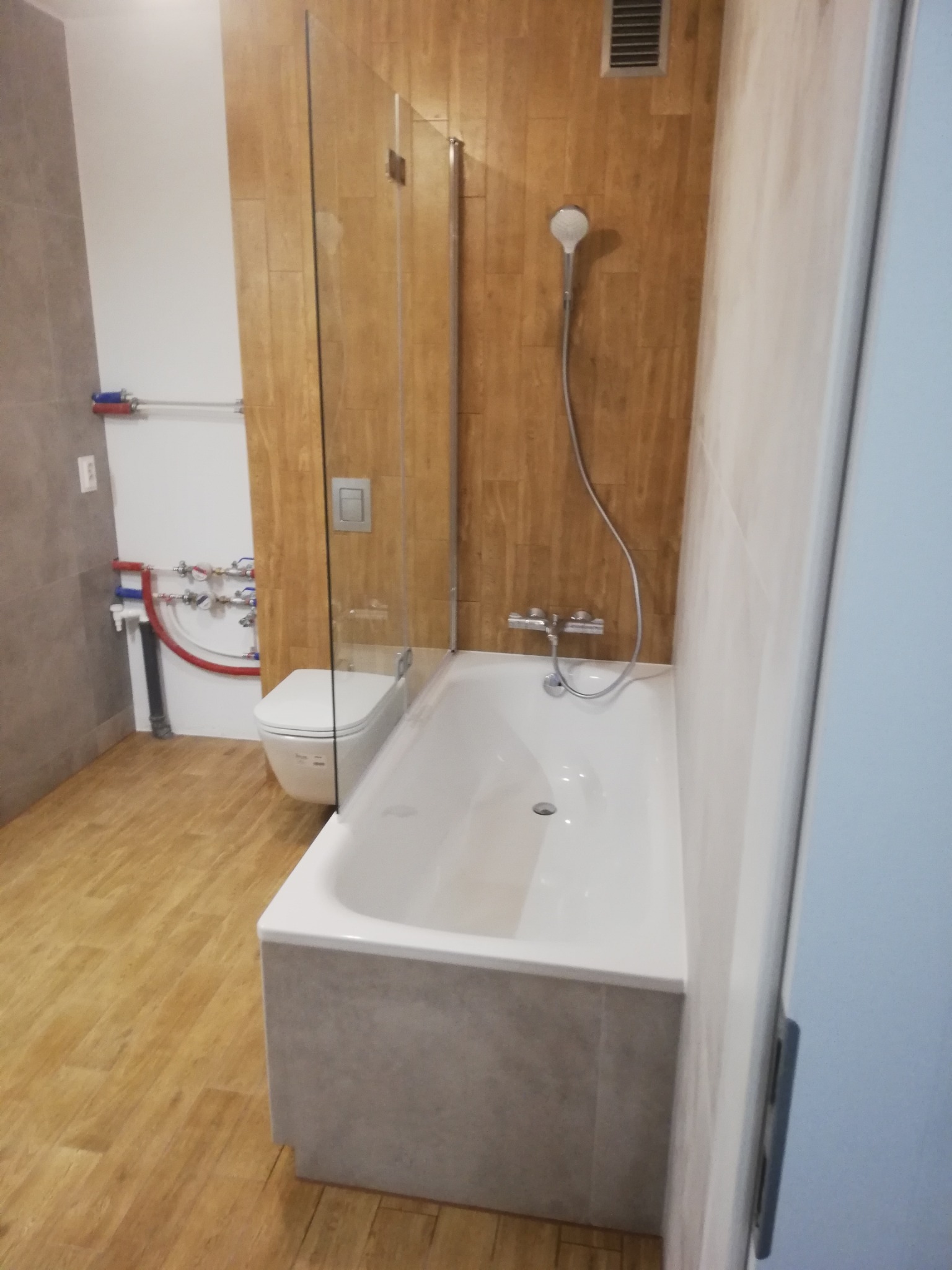 Wykończona łazienka z wanną, szklanym parawanem i toaletą. Drewnopodobne płytki na ścianach i podłodze. Widoczne instalacje wodne i wentylacja.