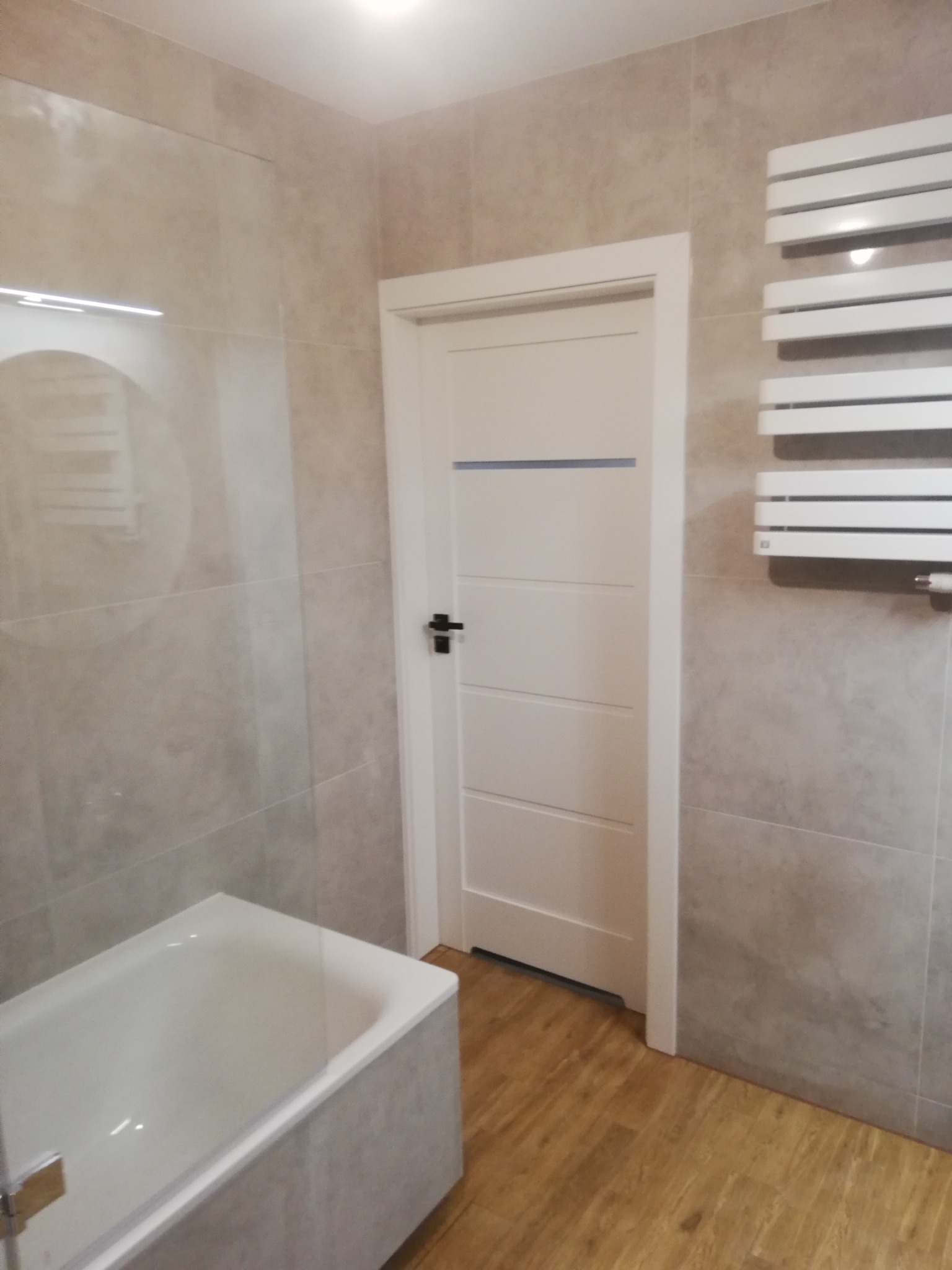 Wykończona łazienka z wanną, grzejnikiem drabinkowym i białą, panelową ścianą. Płytki w odcieniach szarości na ścianach i drewnopodobne na podłodze. Nowoczesny design.