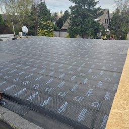 premiumplus - Dach płaski w trakcie krycia papą termozgrzewalną z widocznym logo producenta. W tle zabudowa jednorodzinna i zieleń. Prace dekarskie w toku.