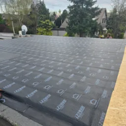 Dach płaski w trakcie krycia papą termozgrzewalną z widocznym logo producenta. W tle zabudowa jednorodzinna i zieleń. Prace dekarskie w toku.