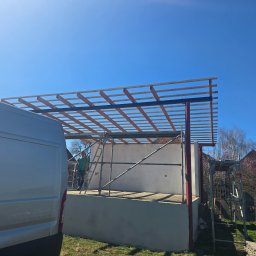 premiumplus - Konstrukcja dachu w trakcie budowy z widocznymi elementami drewnianymi i rusztowaniem, na tle błękitnego nieba i częściowo widocznym samochodem dostawczym.