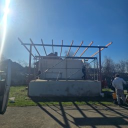 premiumplus - Budowa zadaszenia: konstrukcja z belek na murowanym fundamencie, widoczne rusztowania i pracownicy. Słoneczny dzień, błękitne niebo. Etap budowy.