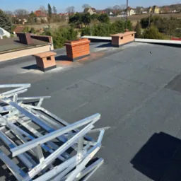 Szary dach płaski z kominem z cegły i elementami wentylacyjnymi. Na pierwszym planie aluminiowa konstrukcja. W tle zabudowa jednorodzinna i drzewa.