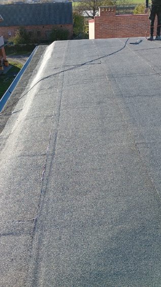 Szary, płaski dach pokryty papą termozgrzewalną, widok z góry. W tle budynek z cegły i fragment zieleni. Widoczny fragment osoby w prawym górnym rogu.