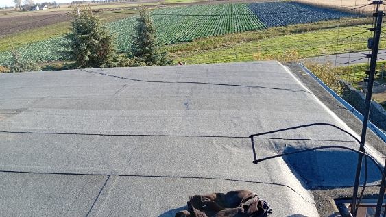 Szary, płaski dach pokryty papą na tle zielonych pól uprawnych. Widoczna antena i fragment odzieży roboczej. Ujęcie z góry, w słoneczny dzień.