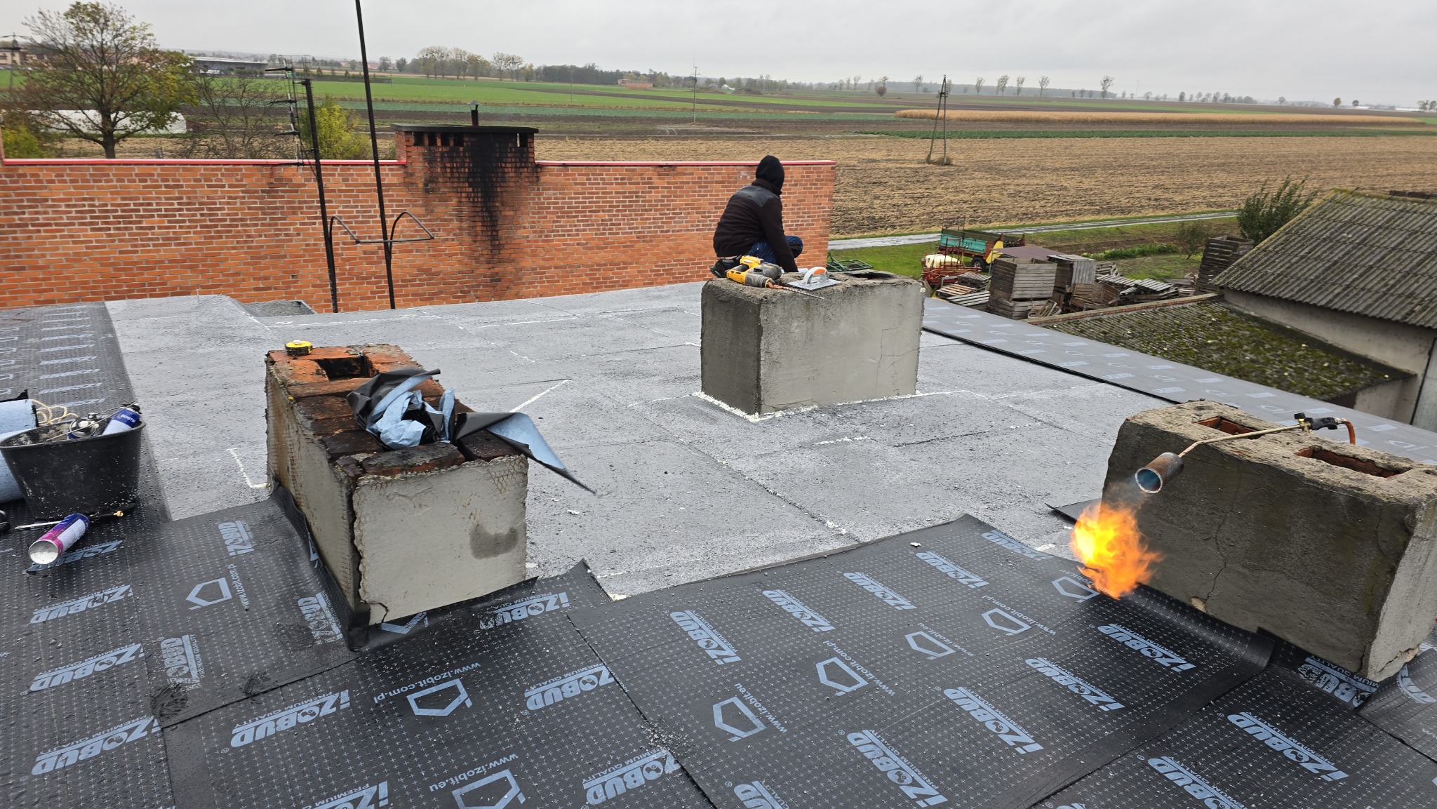 Prace dekarskie na płaskim dachu: papa termozgrzewalna z logo Izobud, palnik dekarski w użyciu, komin. W tle pola uprawne i budynki. Praca w toku, widok z góry.