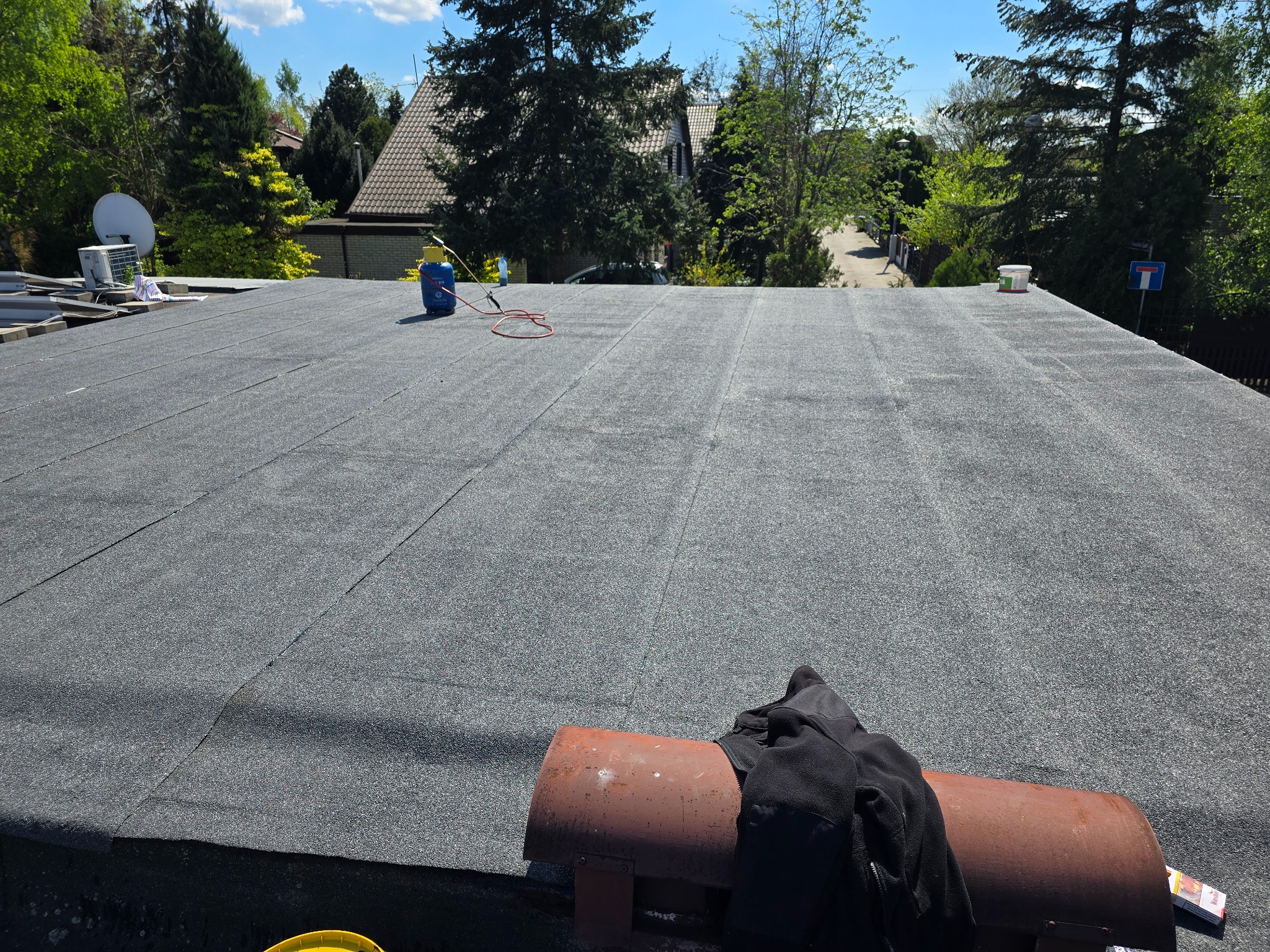 Wykończony, płaski dach pokryty papą termozgrzewalną w słoneczny dzień. Widoczne narzędzia dekarskie i otoczenie z zielenią. Detal: niebieska butla gazowa.