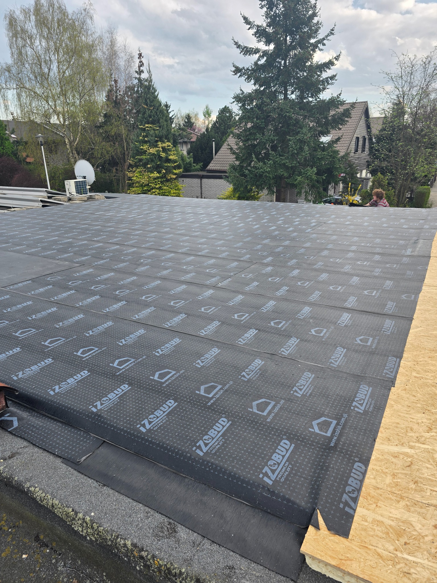 Dach płaski w trakcie krycia papą termozgrzewalną z widocznym logo producenta. W tle zabudowa jednorodzinna i zieleń. Prace dekarskie w toku.