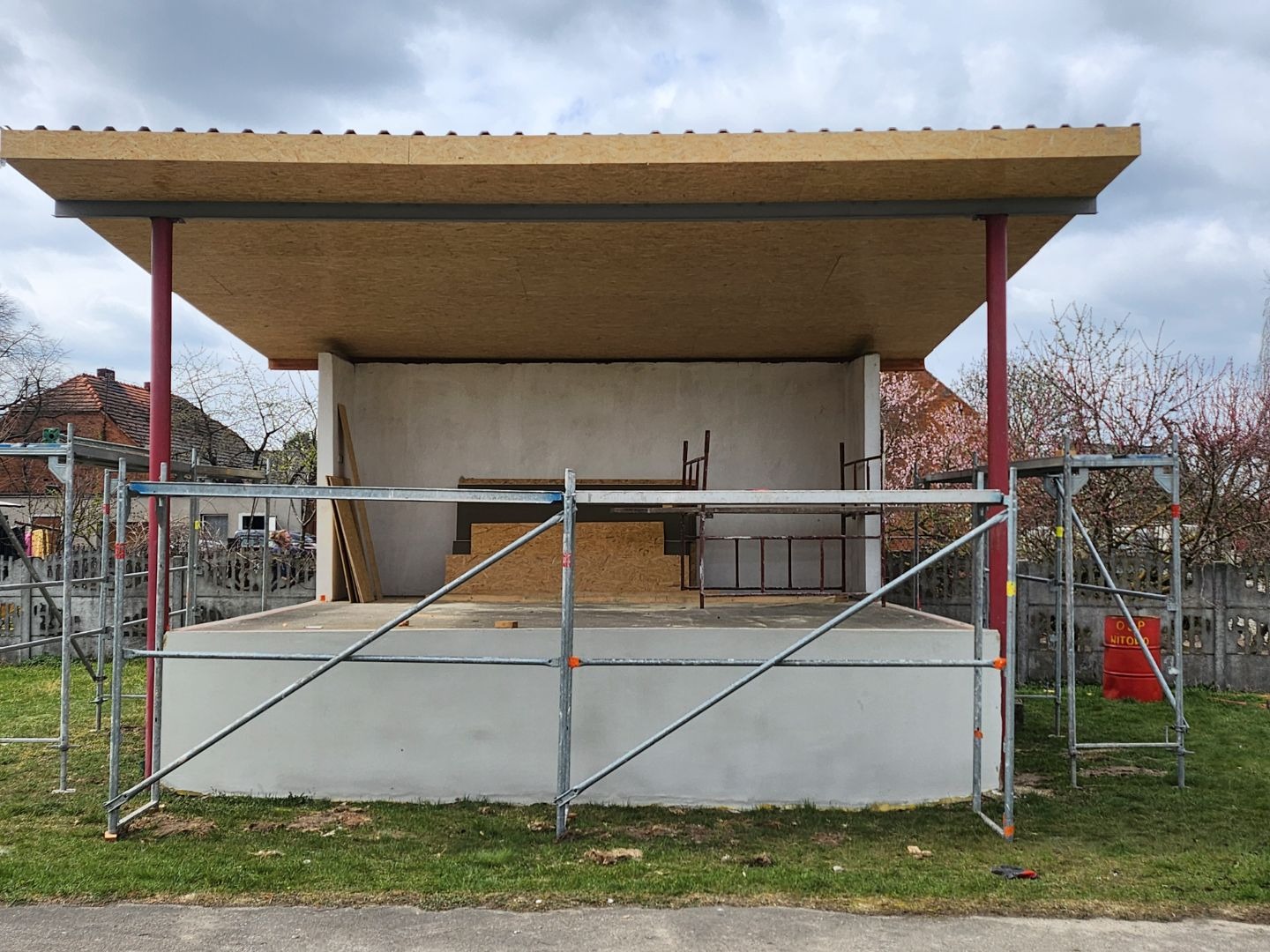 Budowa zadaszonej sceny w plenerze: betonowa podstawa, drewniany dach wsparty na czerwonych kolumnach, rusztowania. Etap prac w słoneczny dzień.