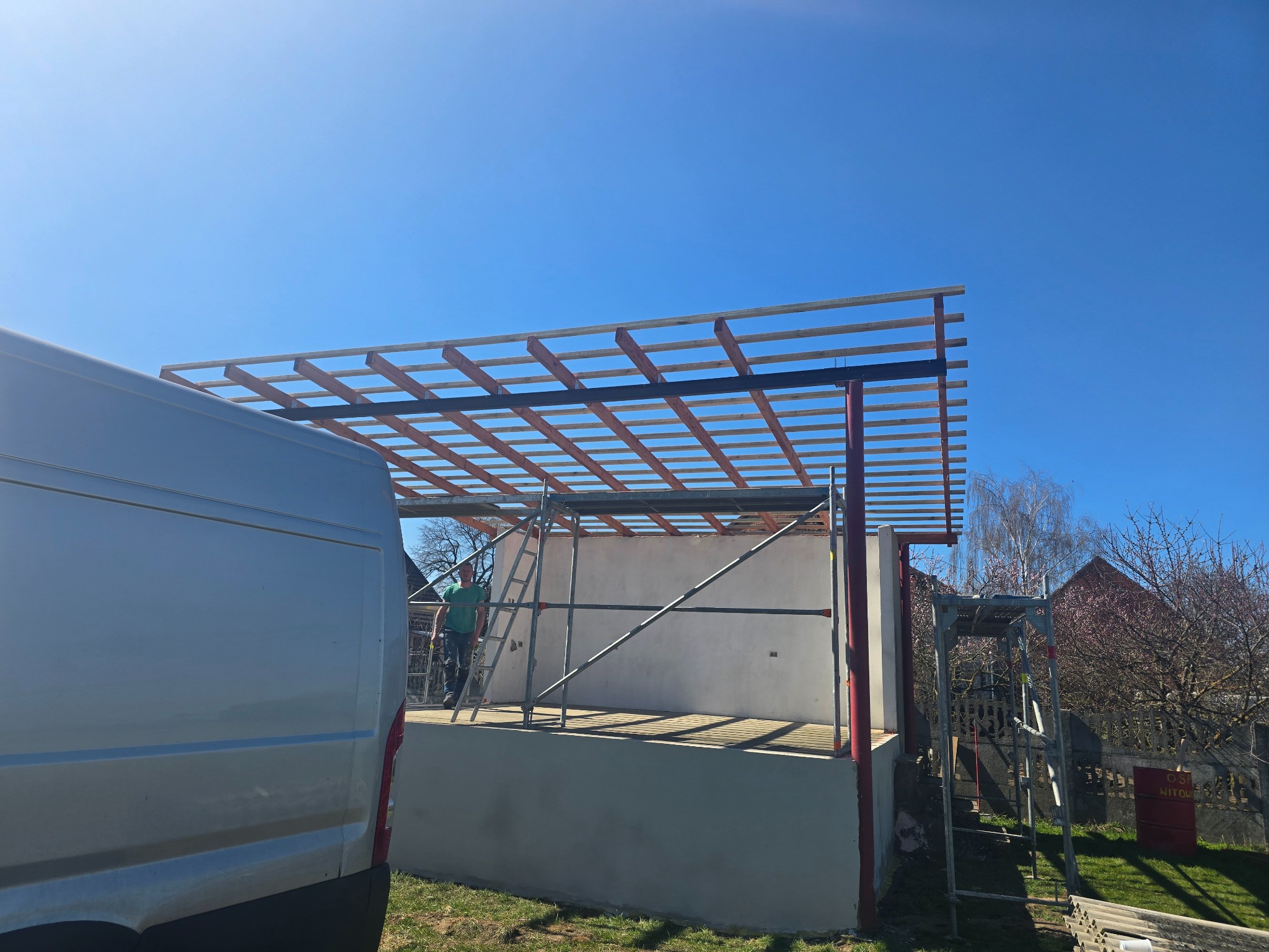 Konstrukcja dachu w trakcie budowy z widocznymi elementami drewnianymi i rusztowaniem, na tle błękitnego nieba i częściowo widocznym samochodem dostawczym.