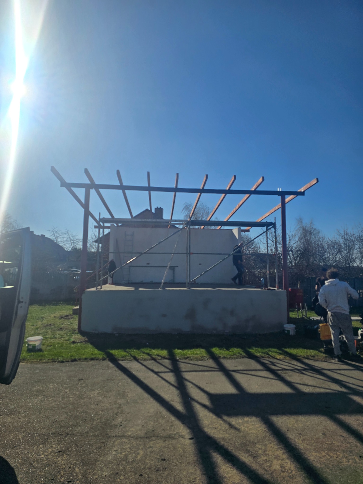 Budowa zadaszenia: konstrukcja z belek na murowanym fundamencie, widoczne rusztowania i pracownicy. Słoneczny dzień, błękitne niebo. Etap budowy.