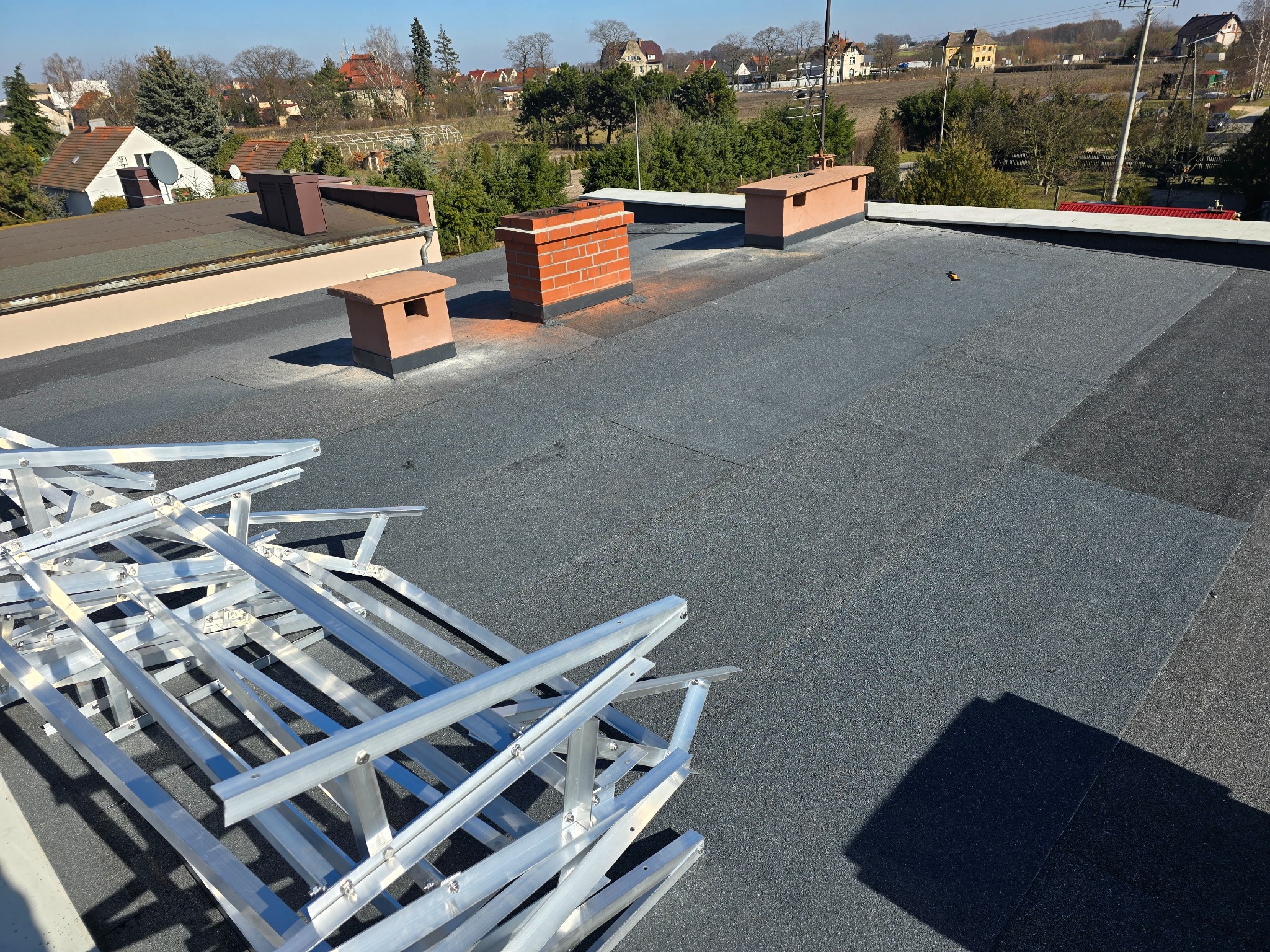 Szary dach płaski z kominem z cegły i elementami wentylacyjnymi. Na pierwszym planie aluminiowa konstrukcja. W tle zabudowa jednorodzinna i drzewa.