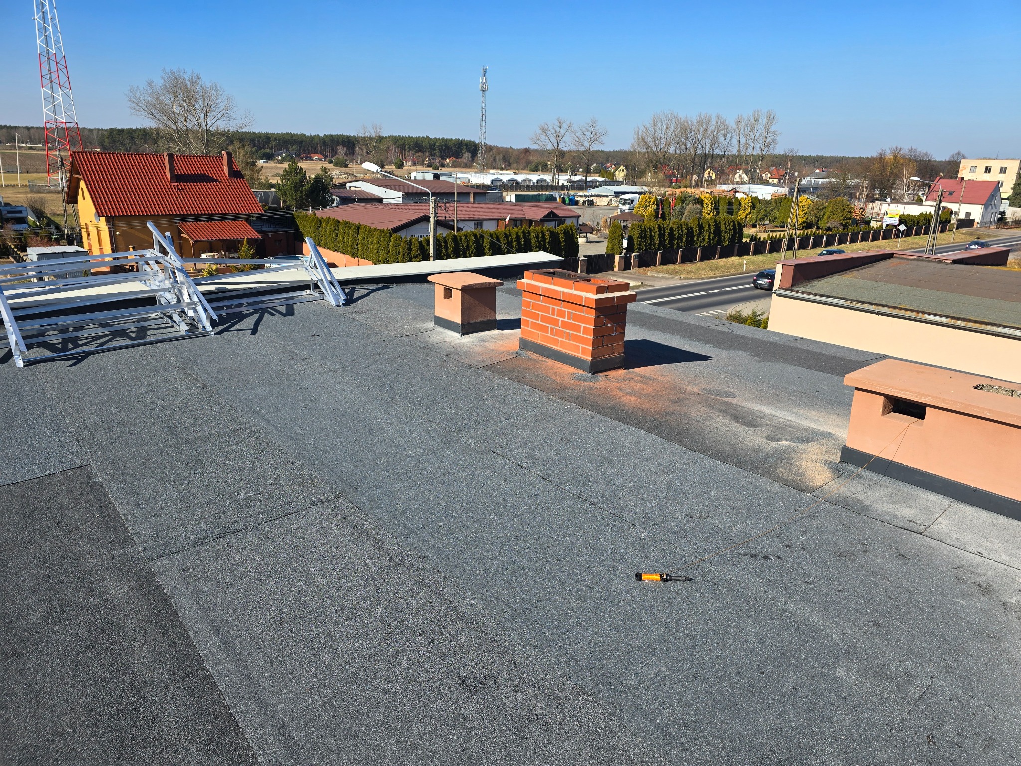 Widok z góry na płaski dach pokryty papą, z kominami i narzędziem dekarskim. W tle zabudowa jednorodzinna i wieża telekomunikacyjna.