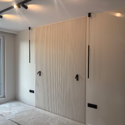 WRESTLING Bohdan Varshavskyi - Ściana z dekoracyjnymi panelami pionowymi w jasnym odcieniu, czarne lampy wiszące i gniazdka, okno z widokiem na zieleń, podłoga zabezpieczona folią malarską.