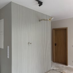 WRESTLING Bohdan Varshavskyi - Ściana z dekoracyjnymi panelami pionowymi w odcieniach szarości, widoczne przygotowania do montażu oświetlenia i drewniane drzwi z czarną klamką. Podłoga zabezpieczona folią.