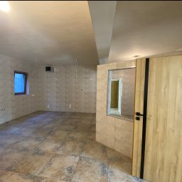 WRESTLING Bohdan Varshavskyi - Wykończone wnętrze z beżowymi płytkami na ścianach i podłodze, widoczne okno i drzwi z czarnymi akcentami. Ściany częściowo wykończone gładzią.