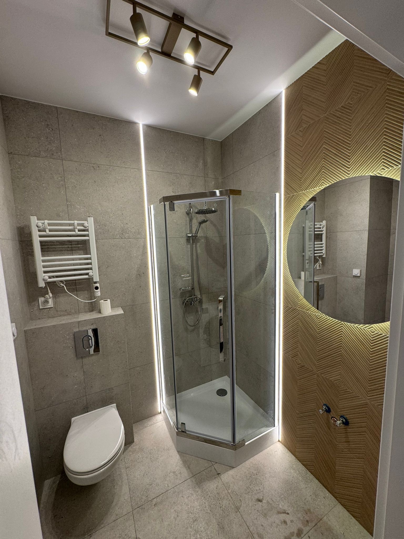 Nowoczesna łazienka z kabiną prysznicową, podświetlanym lustrem w złotej ramie i grzejnikiem drabinkowym. Szare płytki i oświetlenie punktowe tworzą elegancki wystrój.