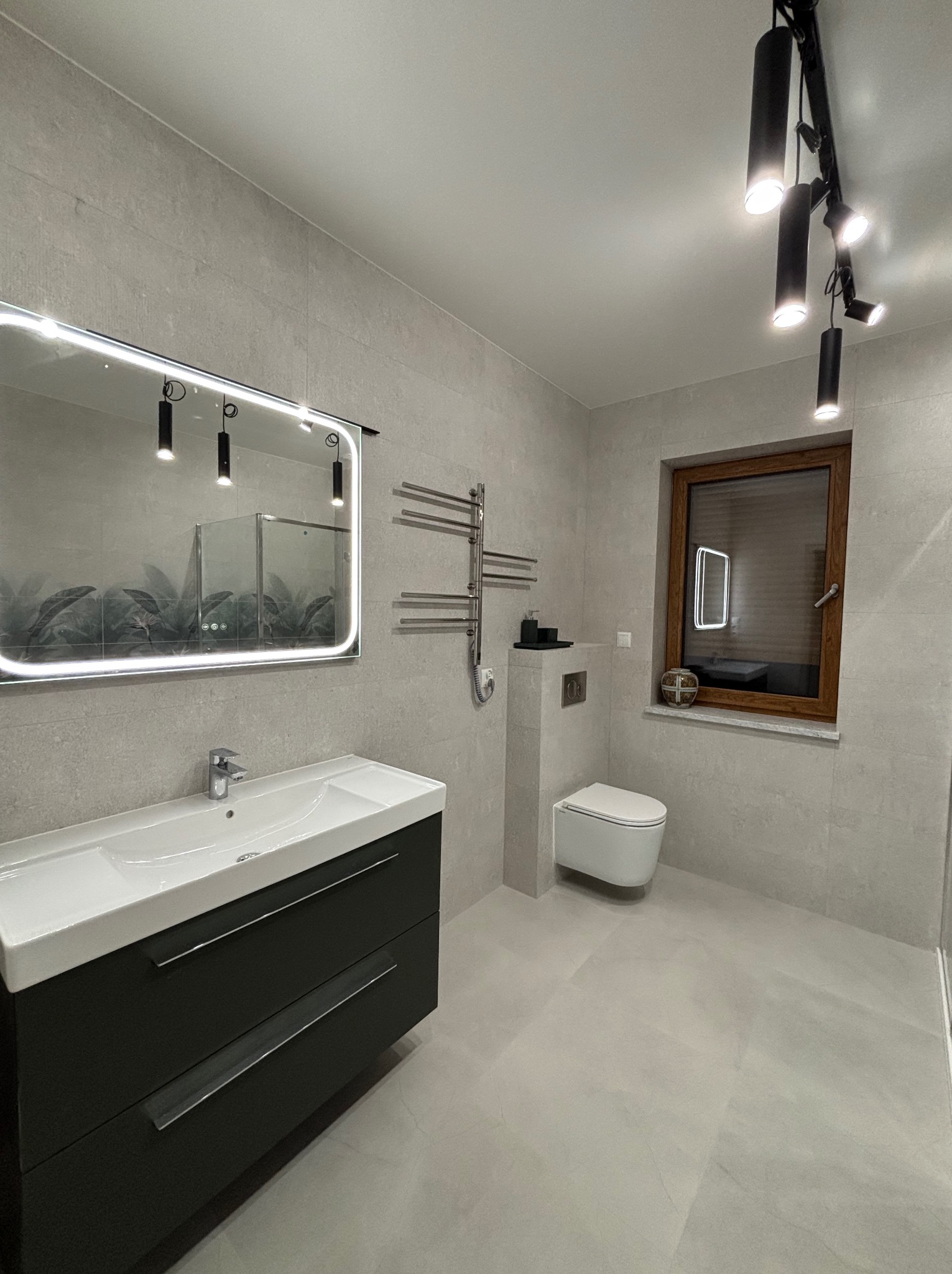 Nowoczesna łazienka w szarościach z podświetlanym lustrem, umywalką z szafką, grzejnikiem drabinkowym i toaletą. Całość dopełnia oświetlenie punktowe.