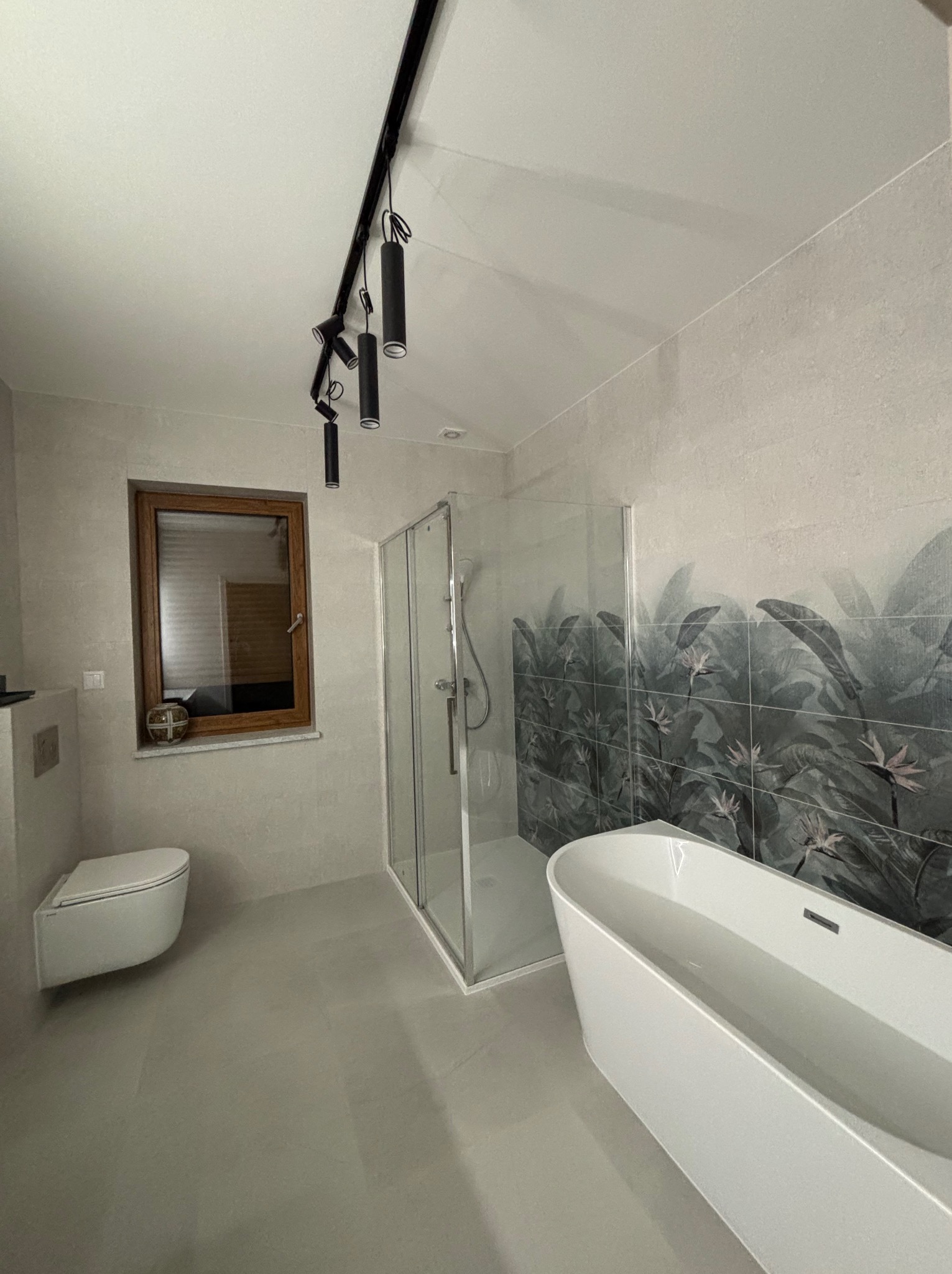 Nowoczesna łazienka w szarościach z akcentem tropikalnym: kabina prysznicowa, wolnostojąca wanna i toaleta podwieszana. Oświetlenie szynowe.