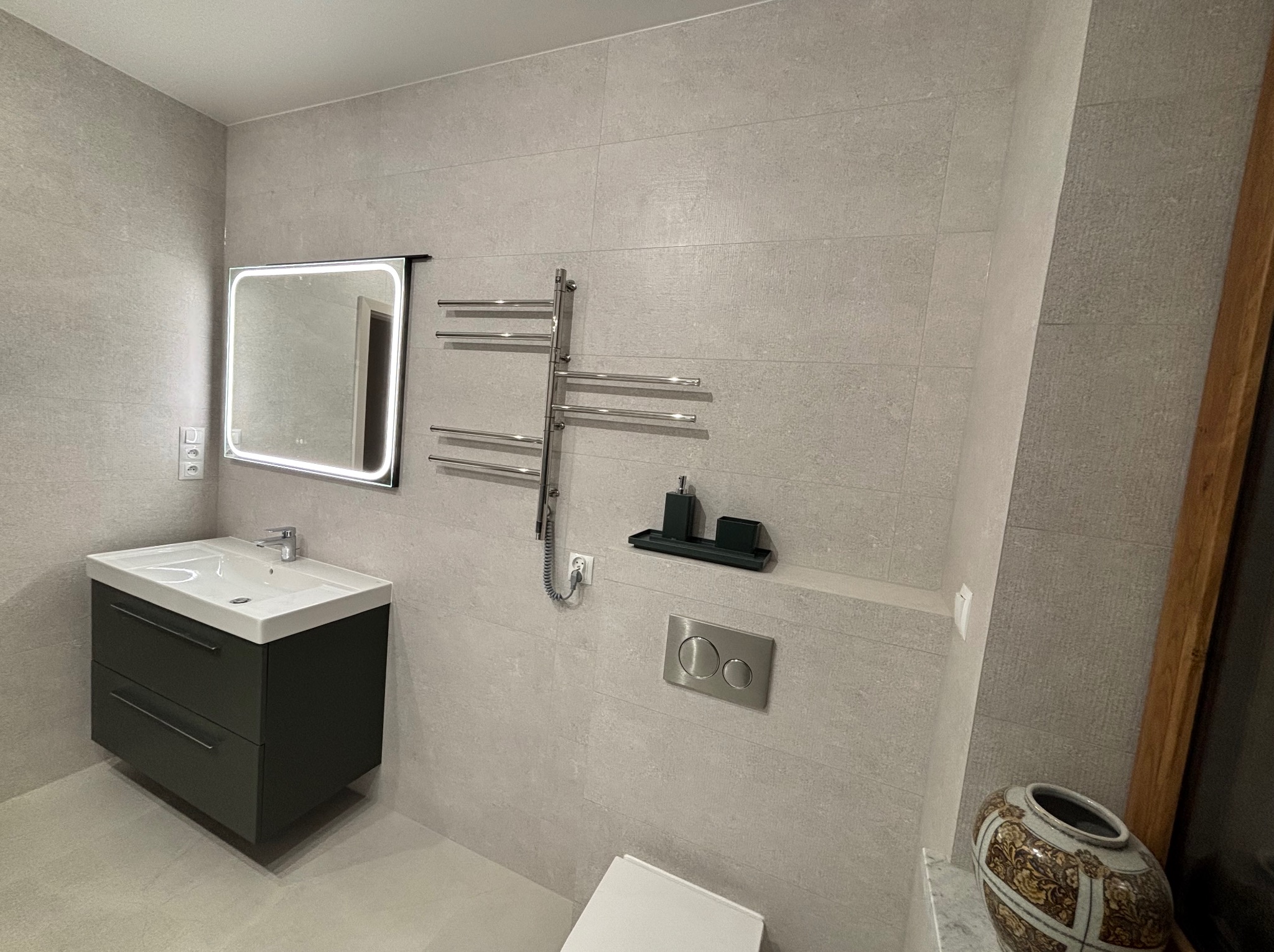 Jasna łazienka po remoncie: szare płytki, podświetlane lustro, grzejnik drabinkowy i umywalka z szafką. Minimalistyczny design z dekoracyjnym wazonem.