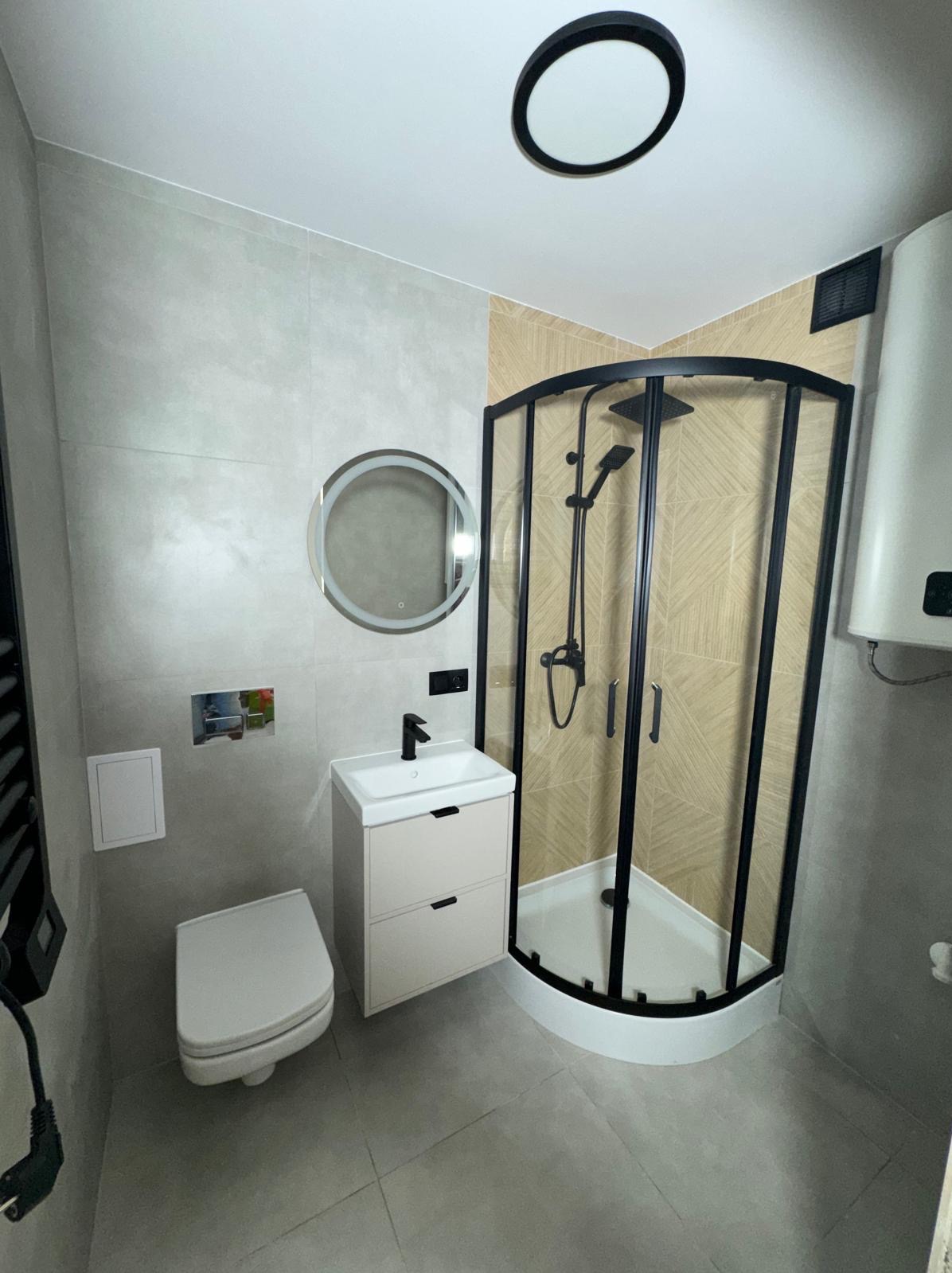 Nowoczesna łazienka po remoncie: kabina prysznicowa z czarną ramą, umywalka z szafką, okrągłe lustro z podświetleniem i szare płytki podłogowe.