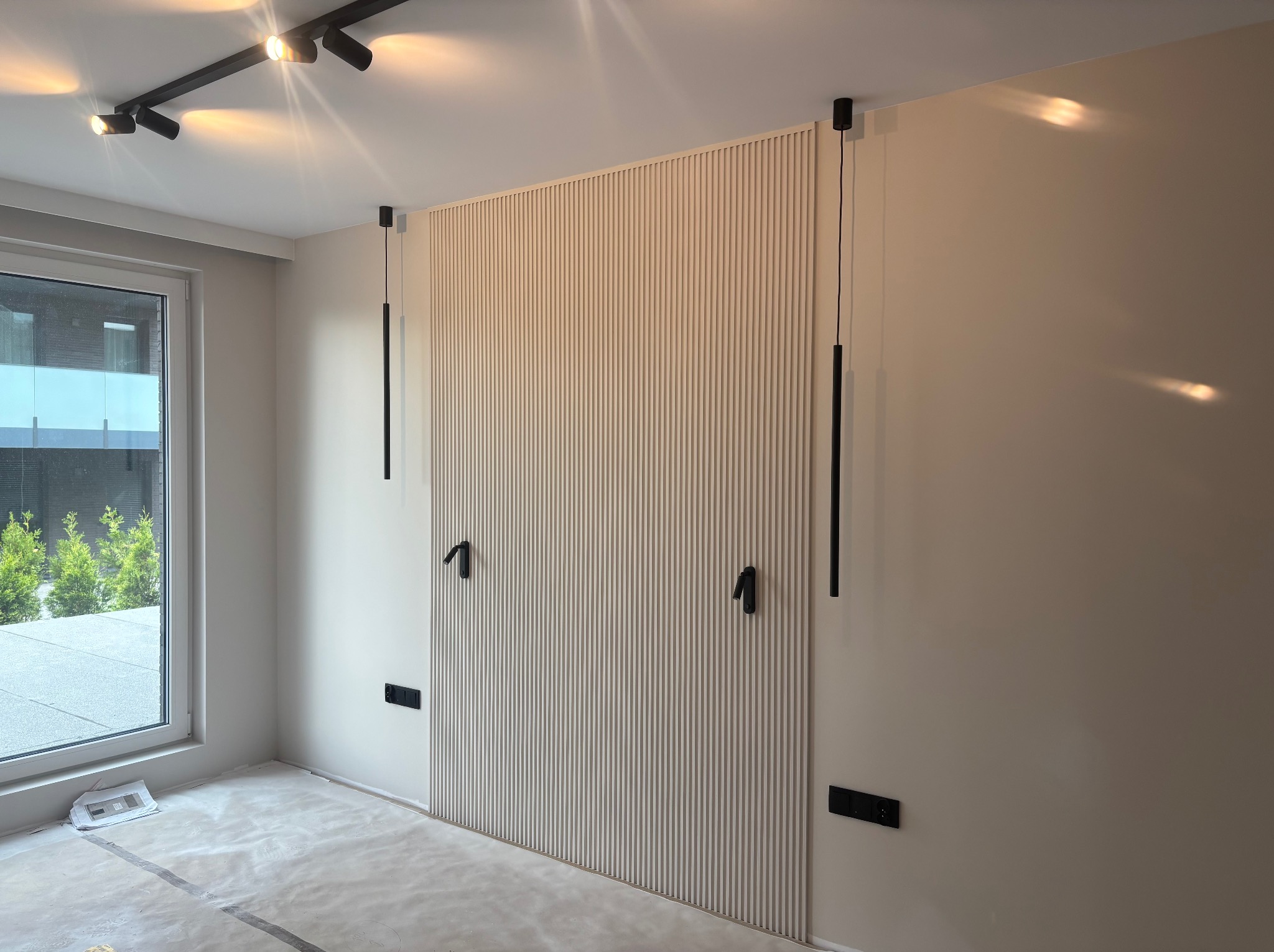 Ściana z dekoracyjnymi panelami pionowymi w jasnym odcieniu, czarne lampy wiszące i gniazdka, okno z widokiem na zieleń, podłoga zabezpieczona folią malarską.