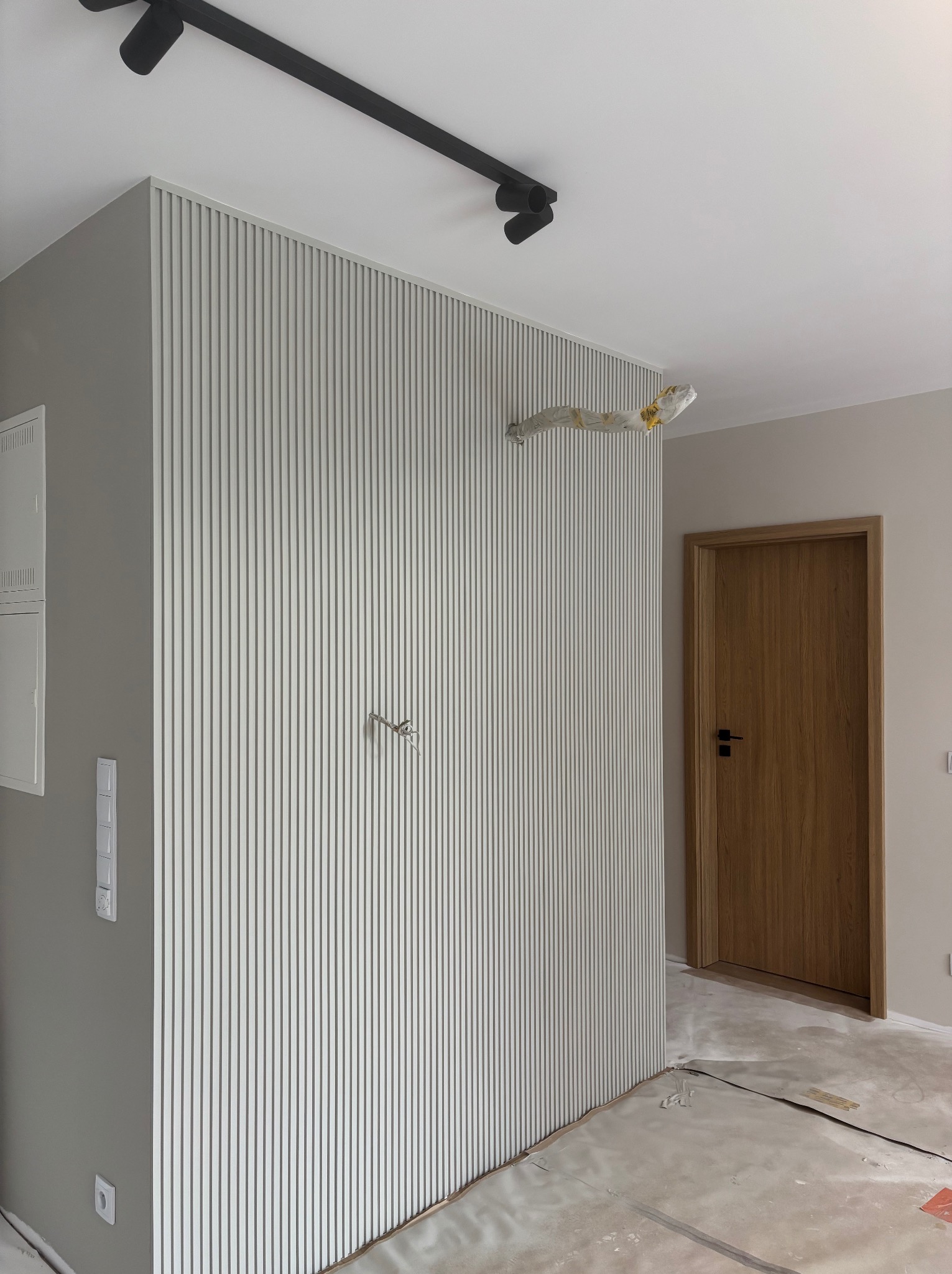 Ściana z dekoracyjnymi panelami pionowymi w odcieniach szarości, widoczne przygotowania do montażu oświetlenia i drewniane drzwi z czarną klamką. Podłoga zabezpieczona folią.