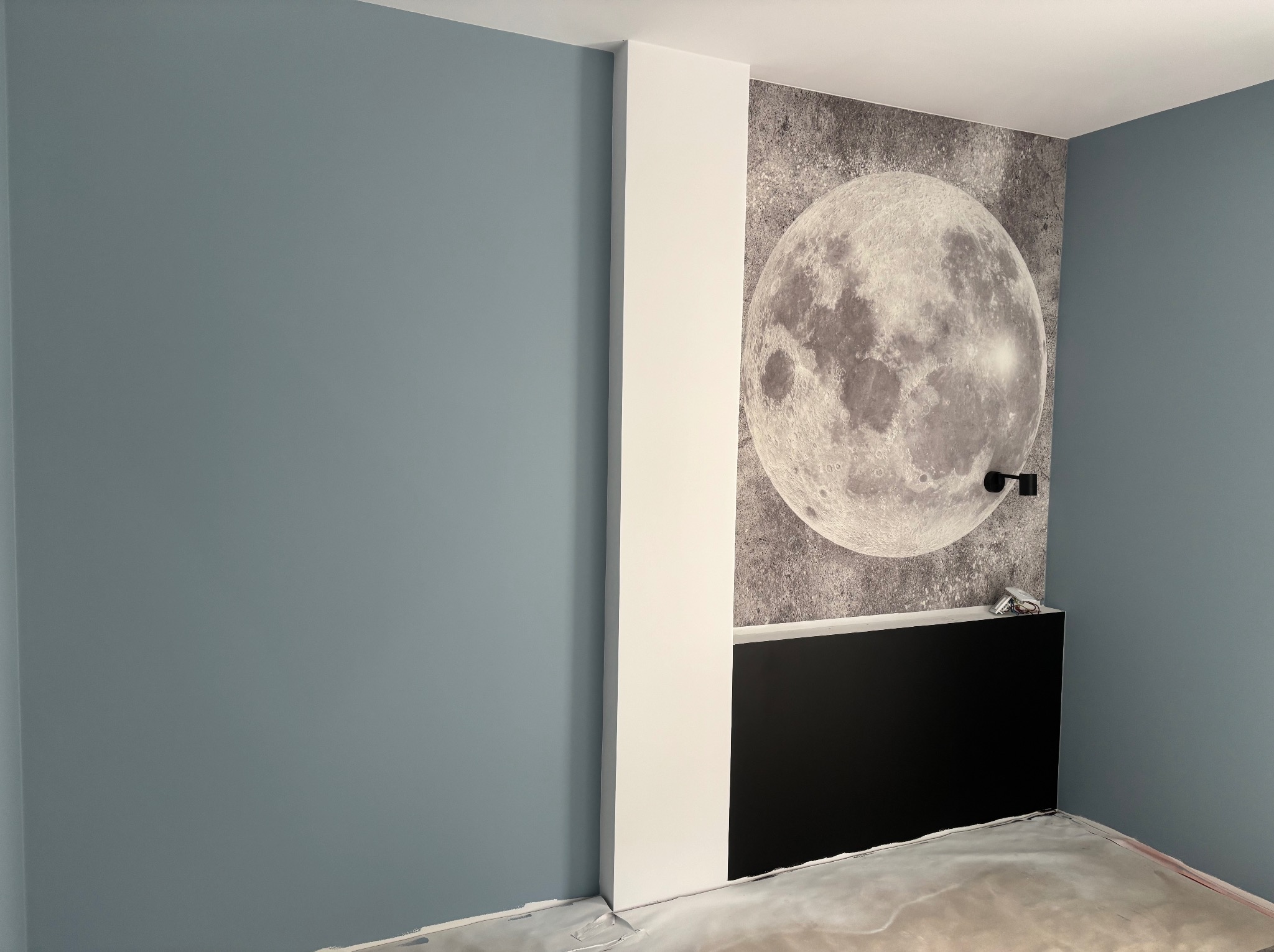 Niebieska ściana, biała kolumna i fototapeta z motywem księżyca w pokoju po remoncie, z widocznymi zabezpieczeniami podłogi.