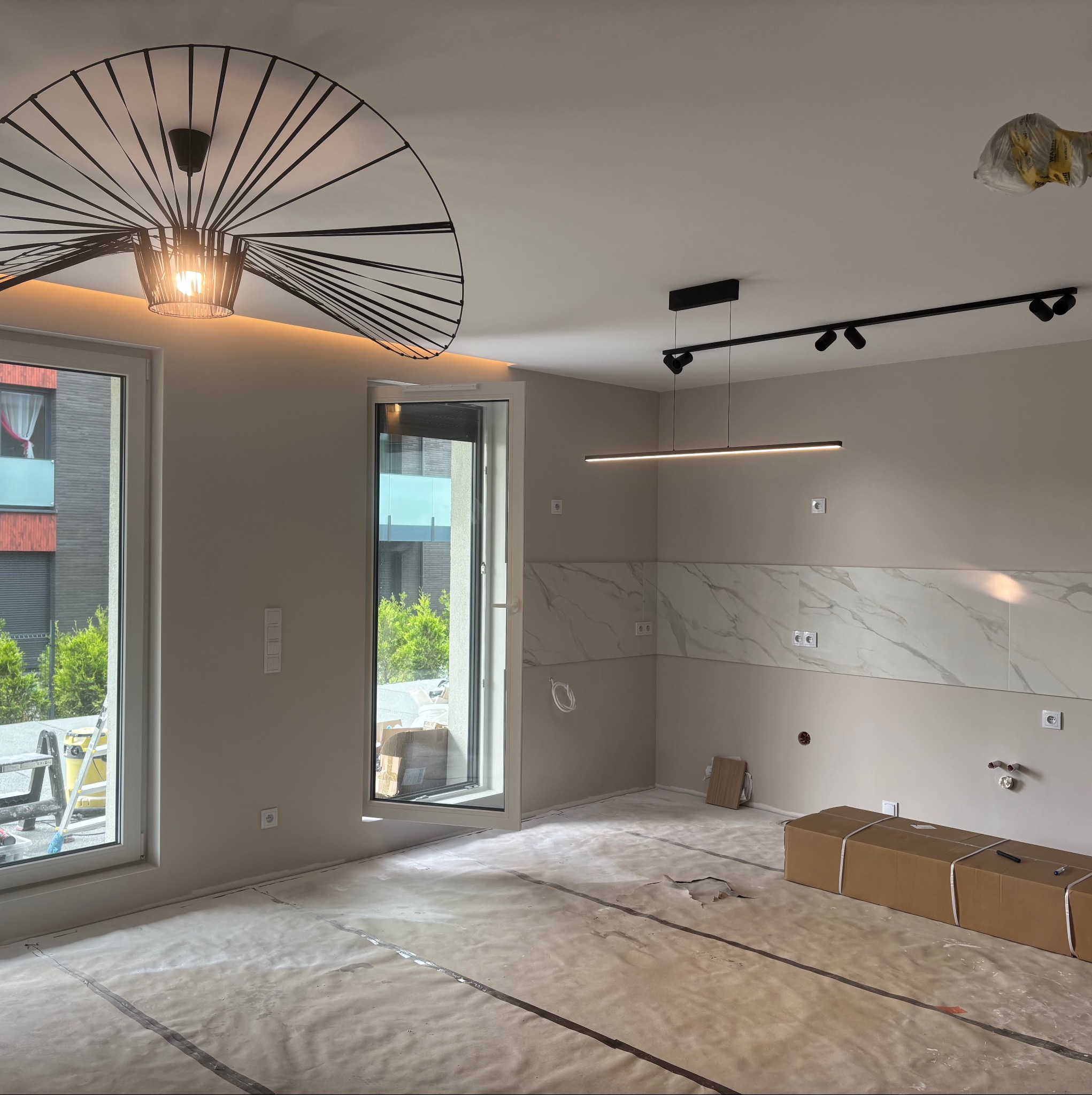 Wnętrze w trakcie wykańczania: zabezpieczona podłoga, fragment ściany z marmurową glazurą, nowoczesne oświetlenie sufitowe. Widoczne okna i elementy instalacji.