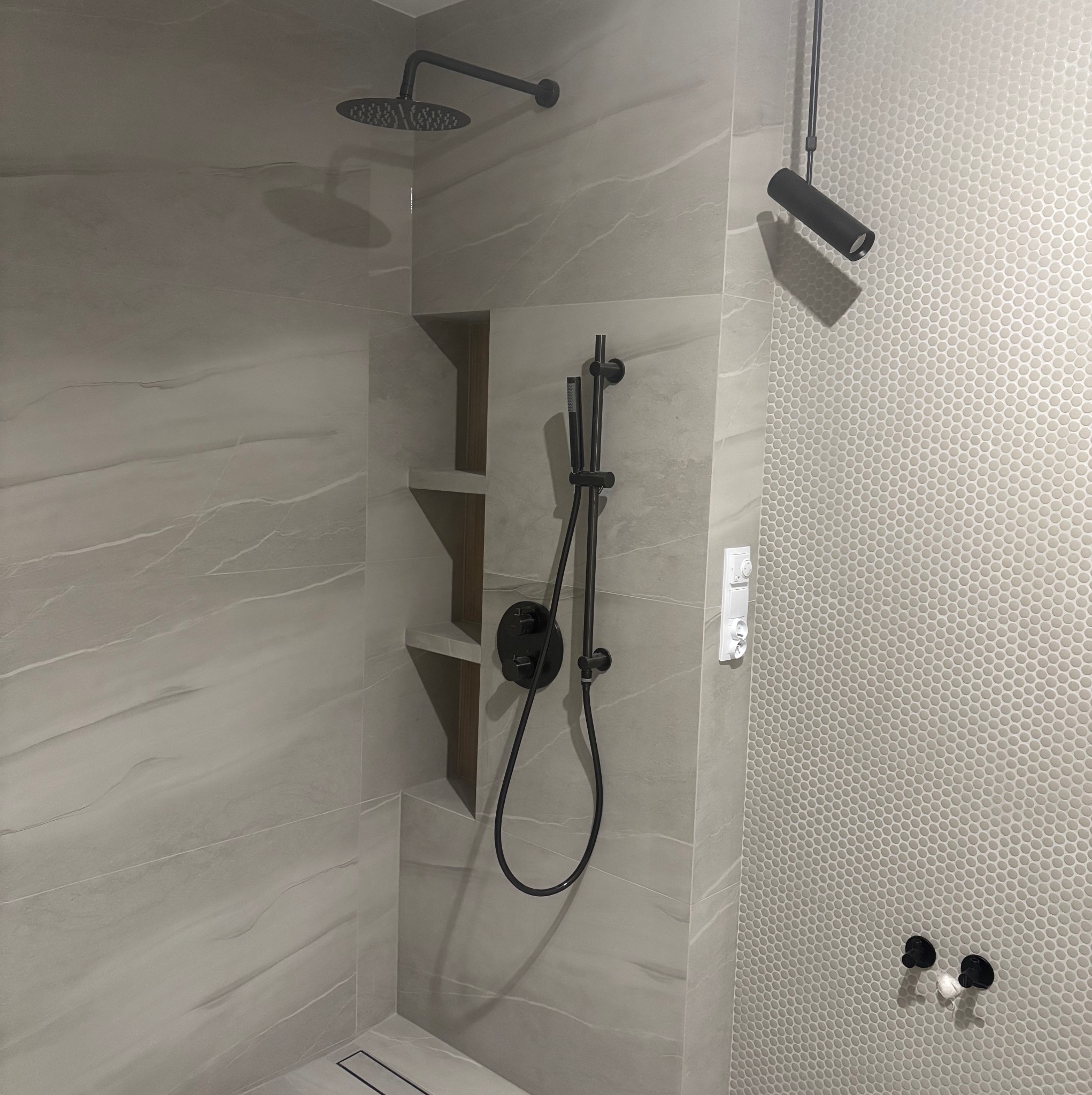 Nowoczesna kabina prysznicowa z szarymi płytkami imitującymi marmur i mozaiką. Czarna armatura, wnęka z półkami i oświetlenie punktowe.