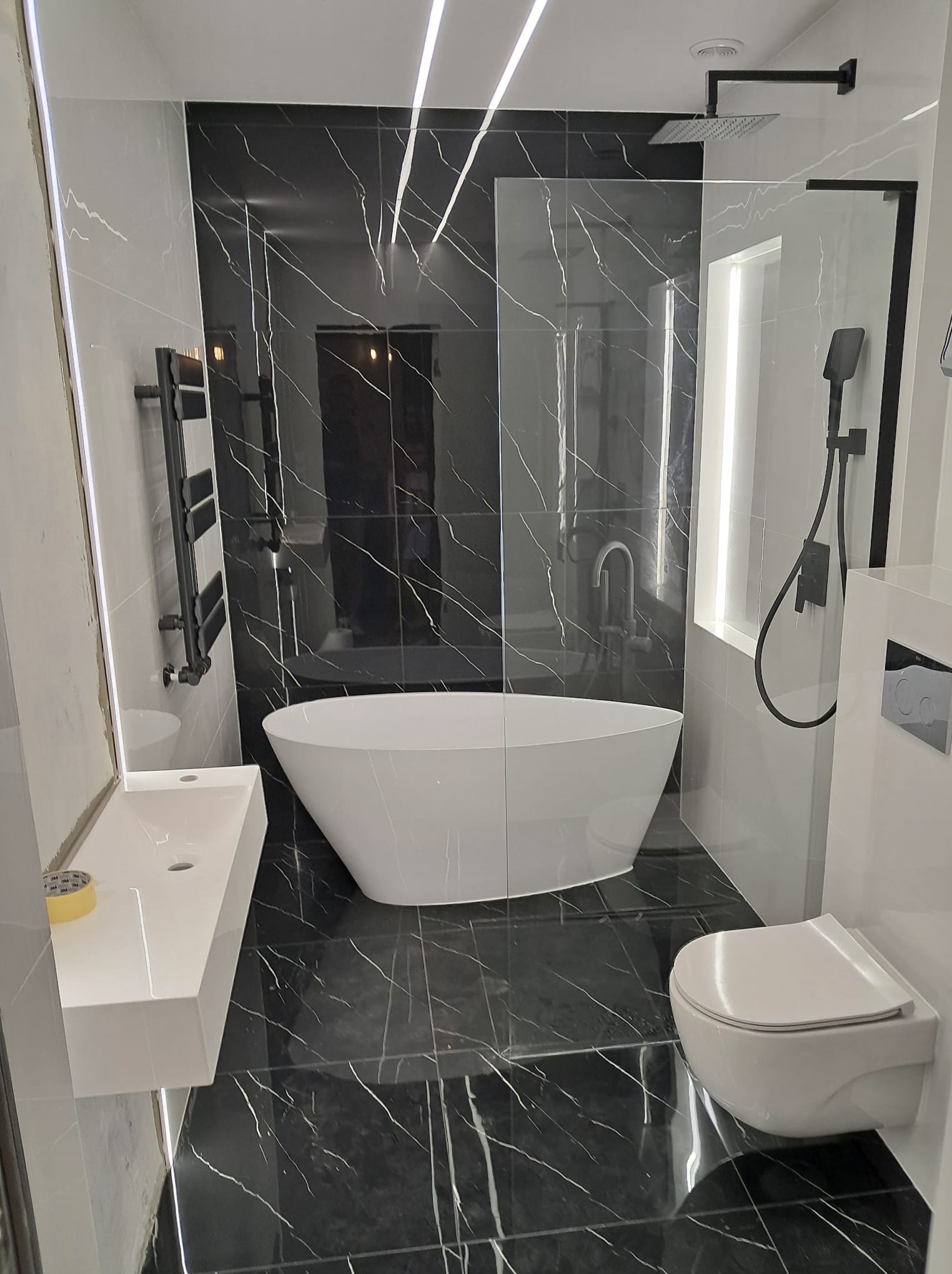 Nowoczesna łazienka z czarno-białymi płytkami imitującymi marmur, wolnostojąca wanna, podświetlane lustro i czarna armatura.