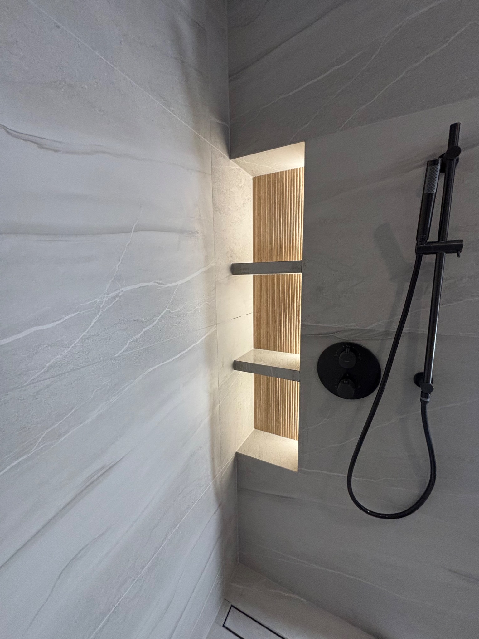 Wykończona łazienka w Gliwicach: szare płytki imitujące marmur, czarna armatura i podświetlana wnęka z drewnianym akcentem i półkami.