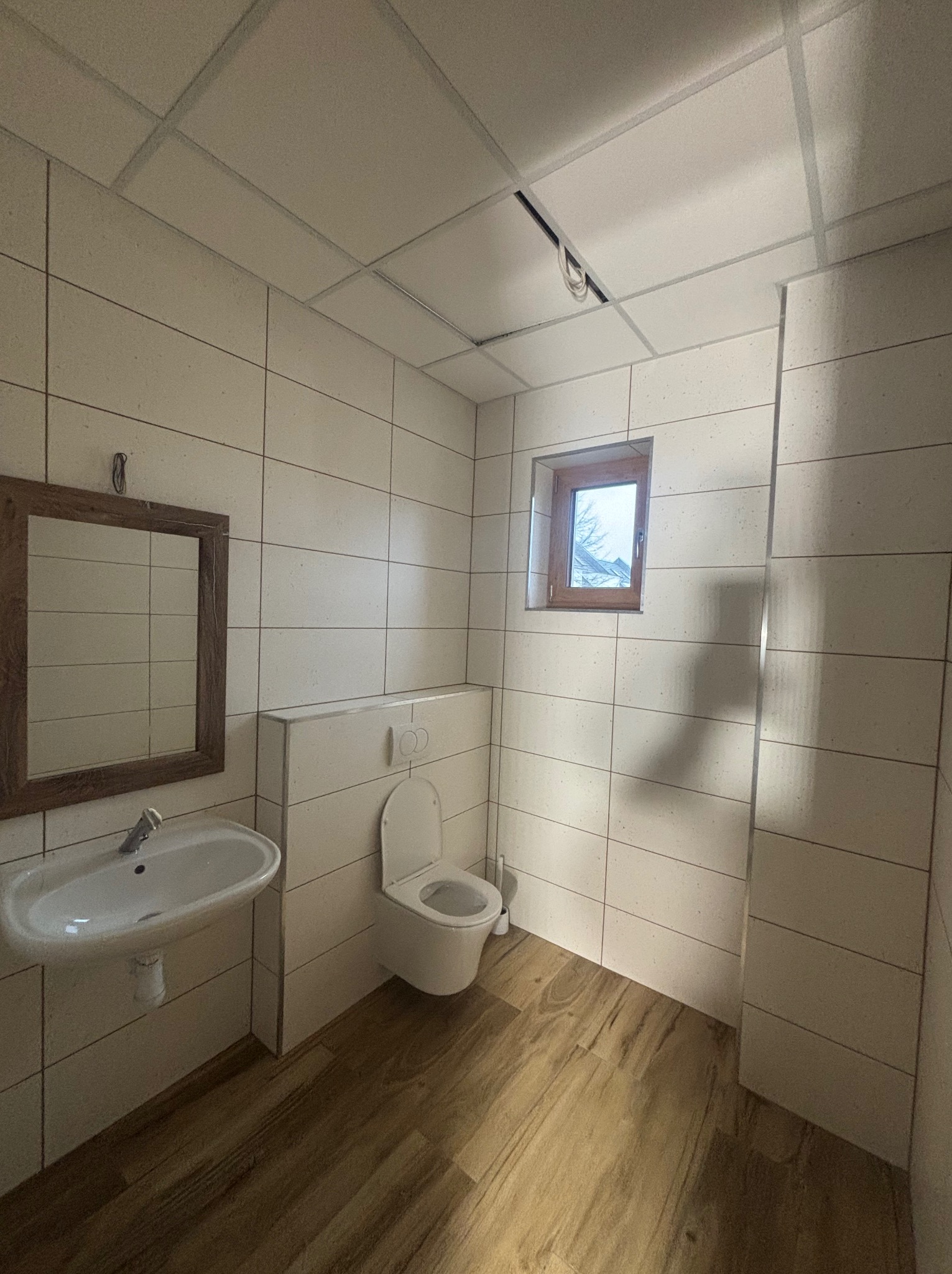 Wykończona łazienka z beżowymi płytkami, drewnianą podłogą i oknem. Umywalka z lustrem po lewej, toaleta po prawej. Sufit podwieszany z oświetleniem.