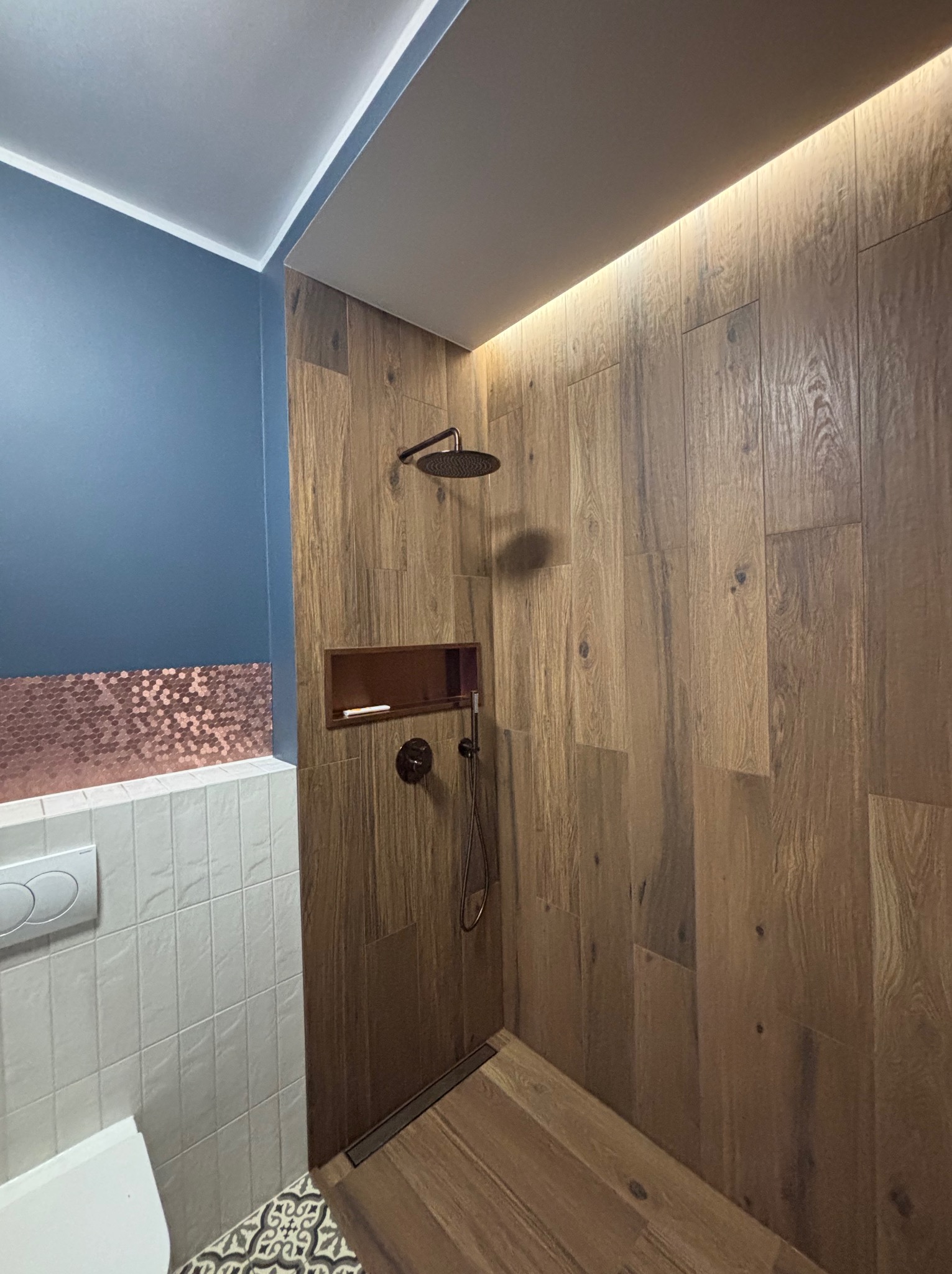 Nowoczesna kabina prysznicowa z drewnopodobnymi płytkami, oświetleniem LED i elementami w kolorze miedzi. Widoczna armatura w ciemnym kolorze i dekoracyjna mozaika podłogowa.