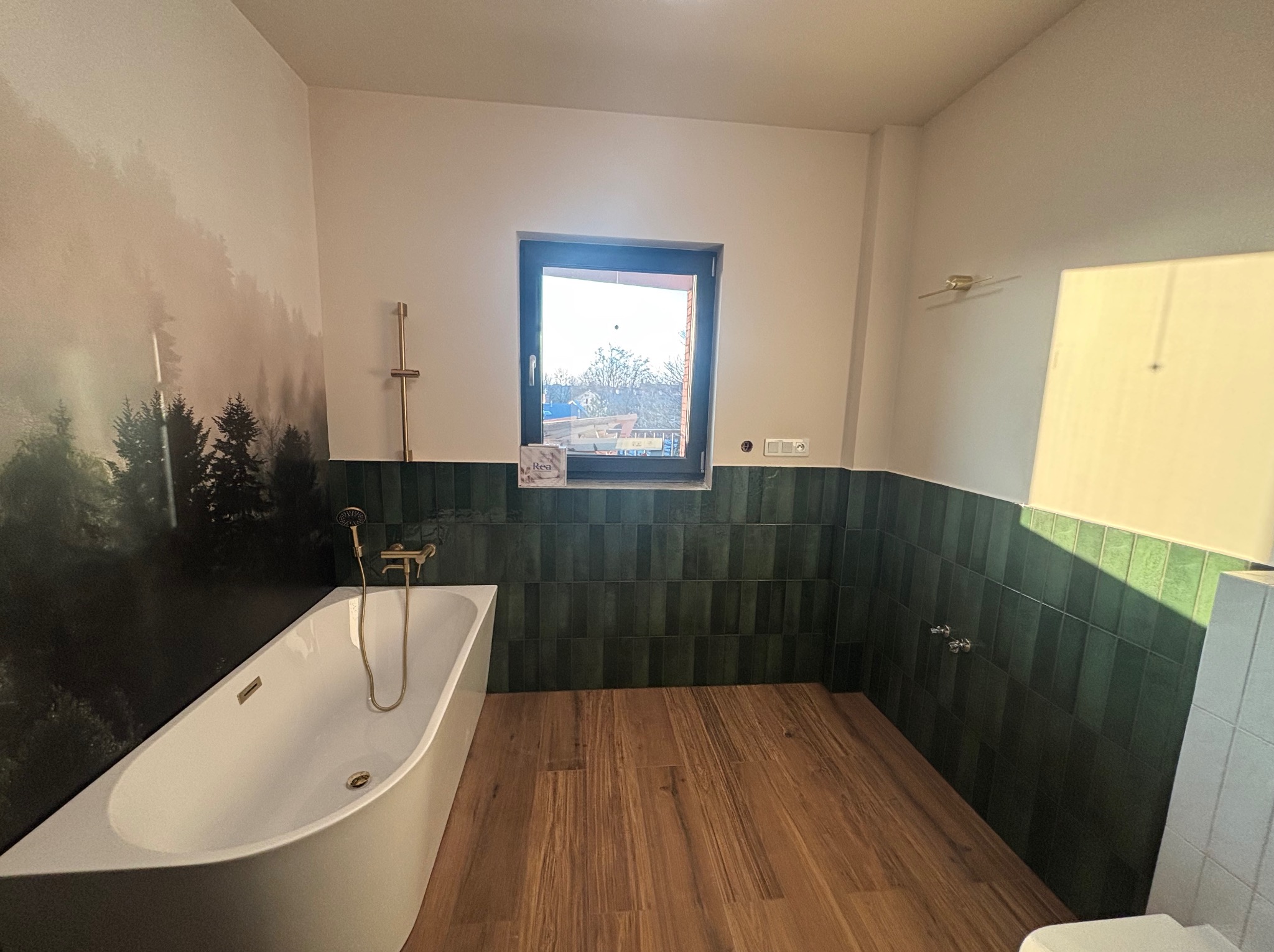 Nowoczesna łazienka z wanną, zielonymi płytkami i drewnianą podłogą. Na ścianie fototapeta z motywem lasu. Widoczne okno z widokiem na zewnątrz.